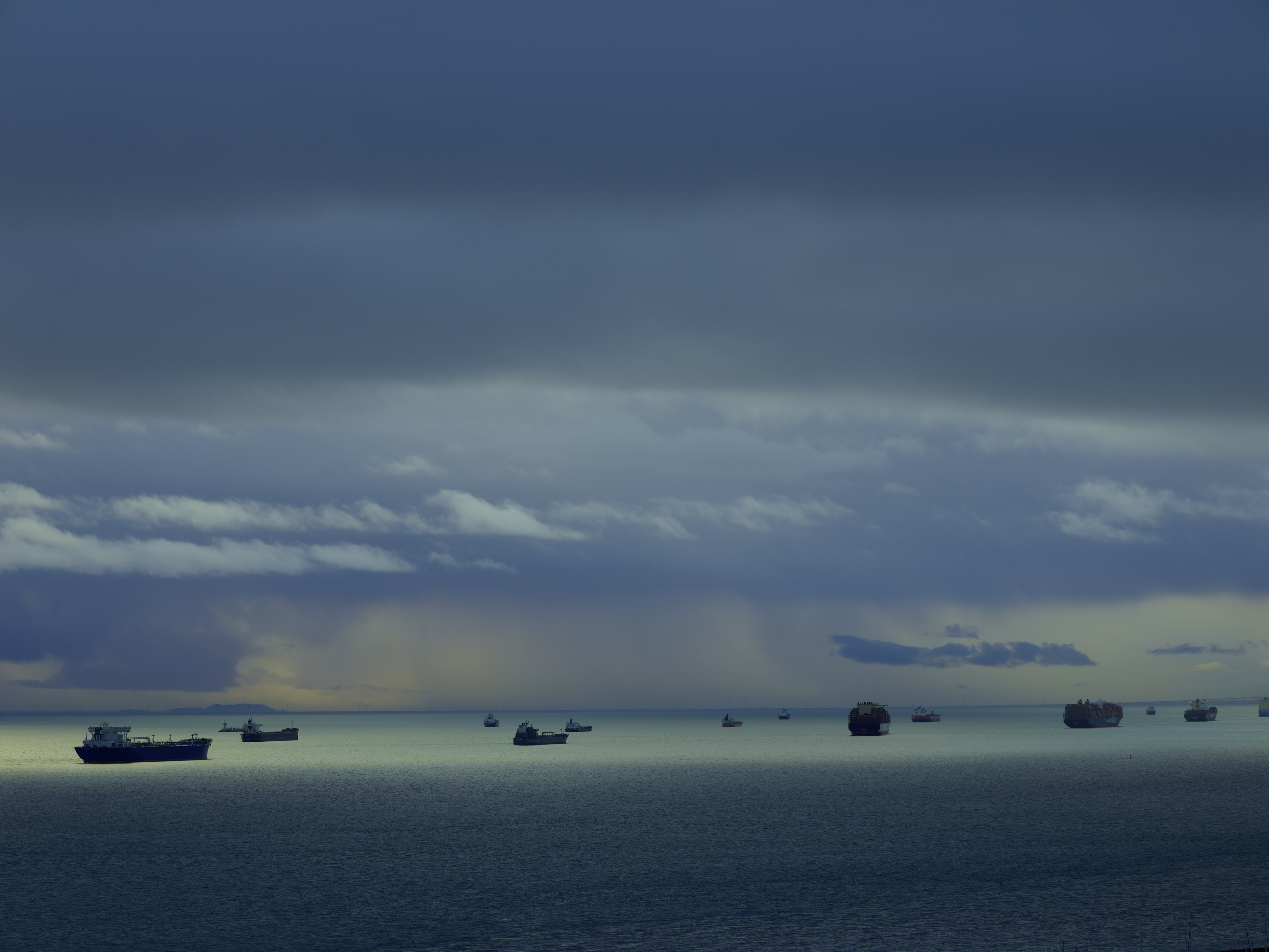 Ships silhouetted on dark blue water beneath cloudy grey sky with yellow-green tinged clouds on horizon.