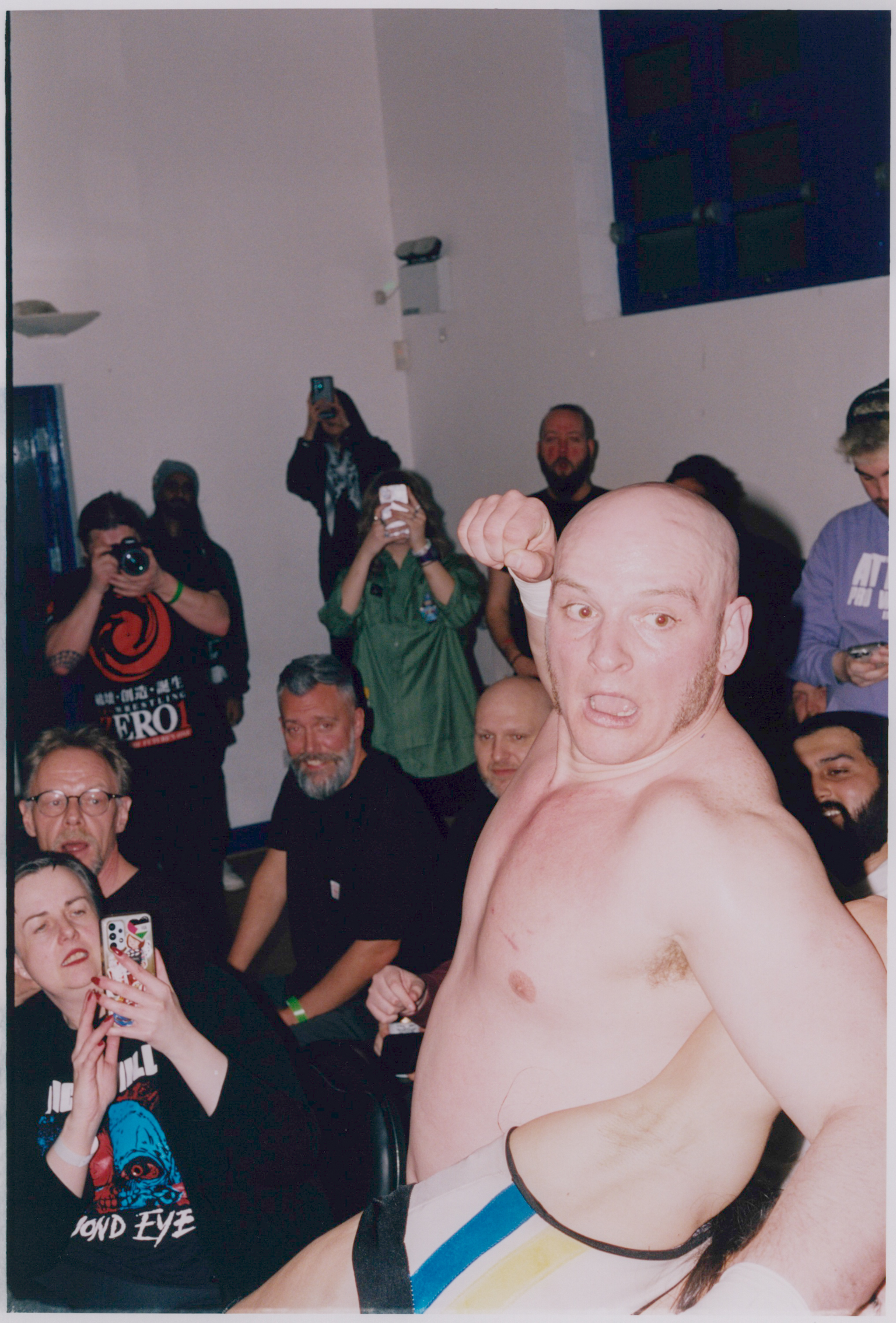 Shirtless bald man in foreground with crowd of onlookers behind him in white-walled room, some holding phones and cameras.