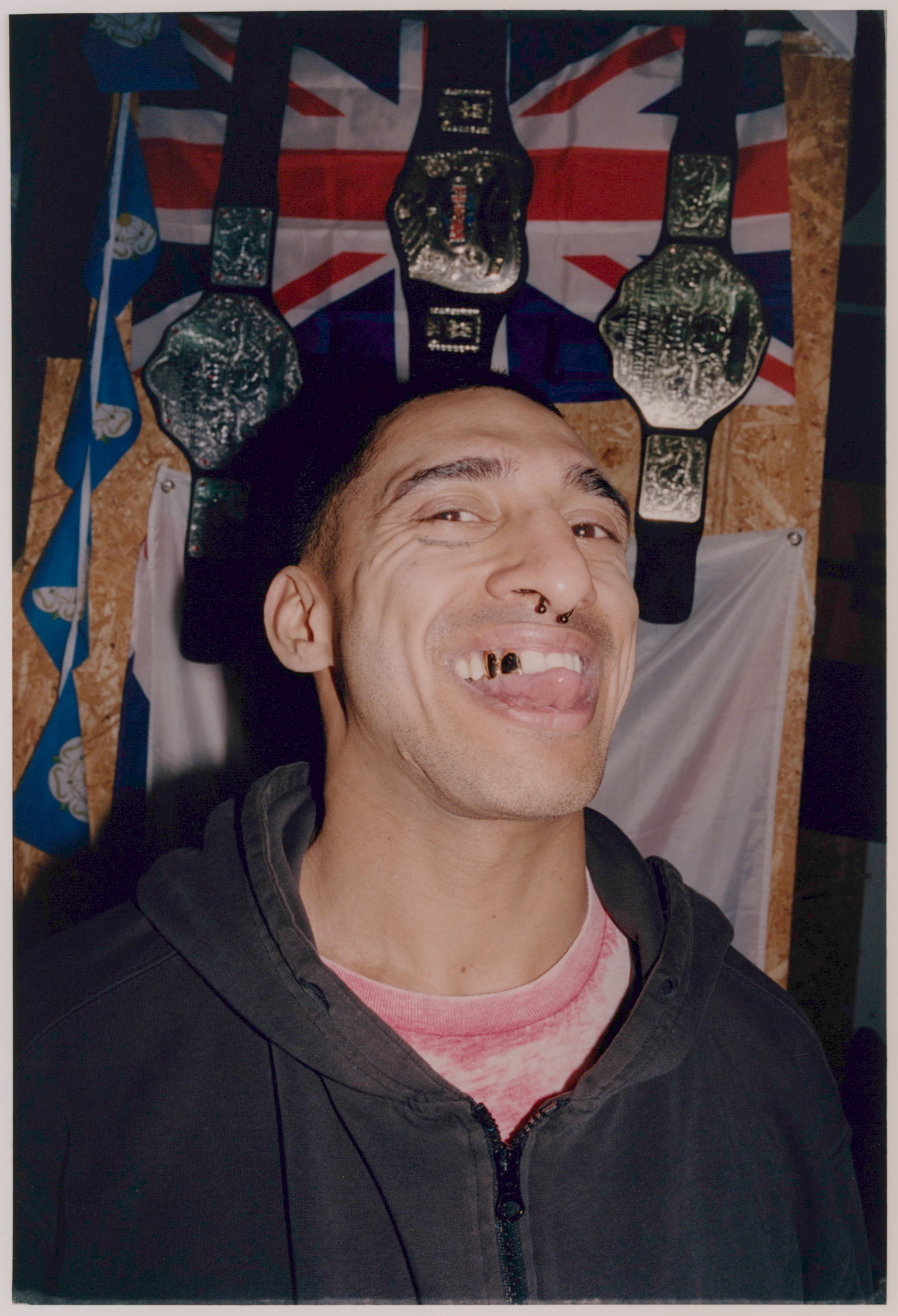 Man in dark hoodie smiling with missing teeth, standing before Union Jack flag and championship belts on wall.