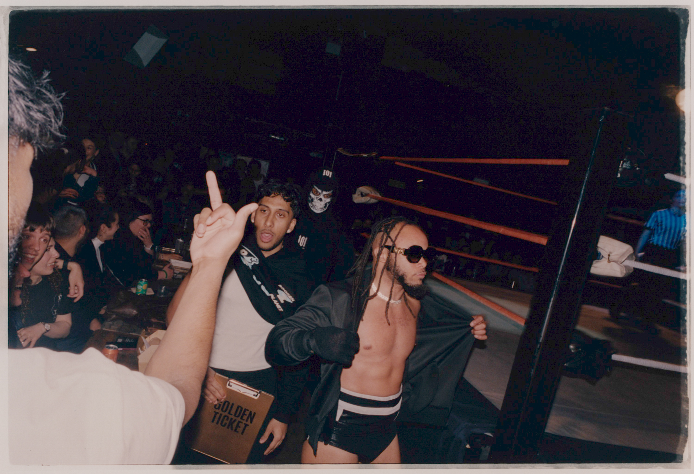 Person with raised finger gesture in crowded indoor space with stairs and dim lighting. Multiple people visible in background.