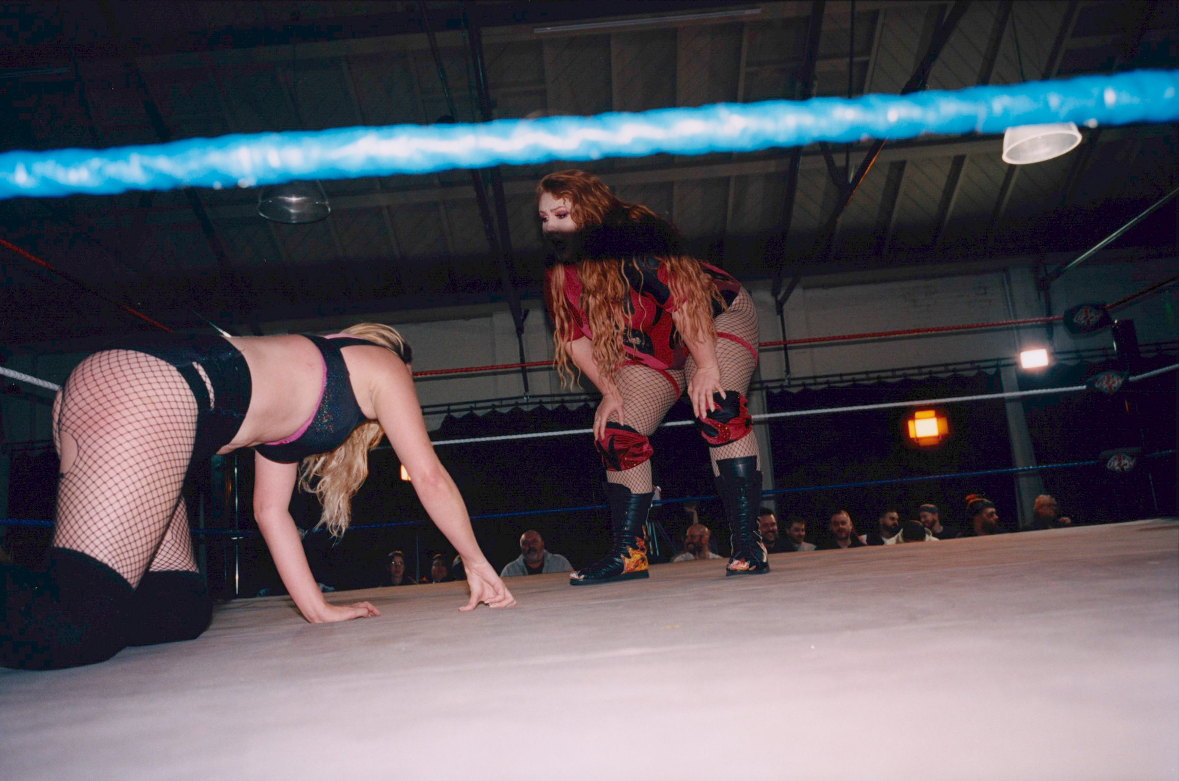 Two wrestlers in a ring, one in fishnet stockings crouched low, another in red and gold mask standing behind. Blue lighting overhead.