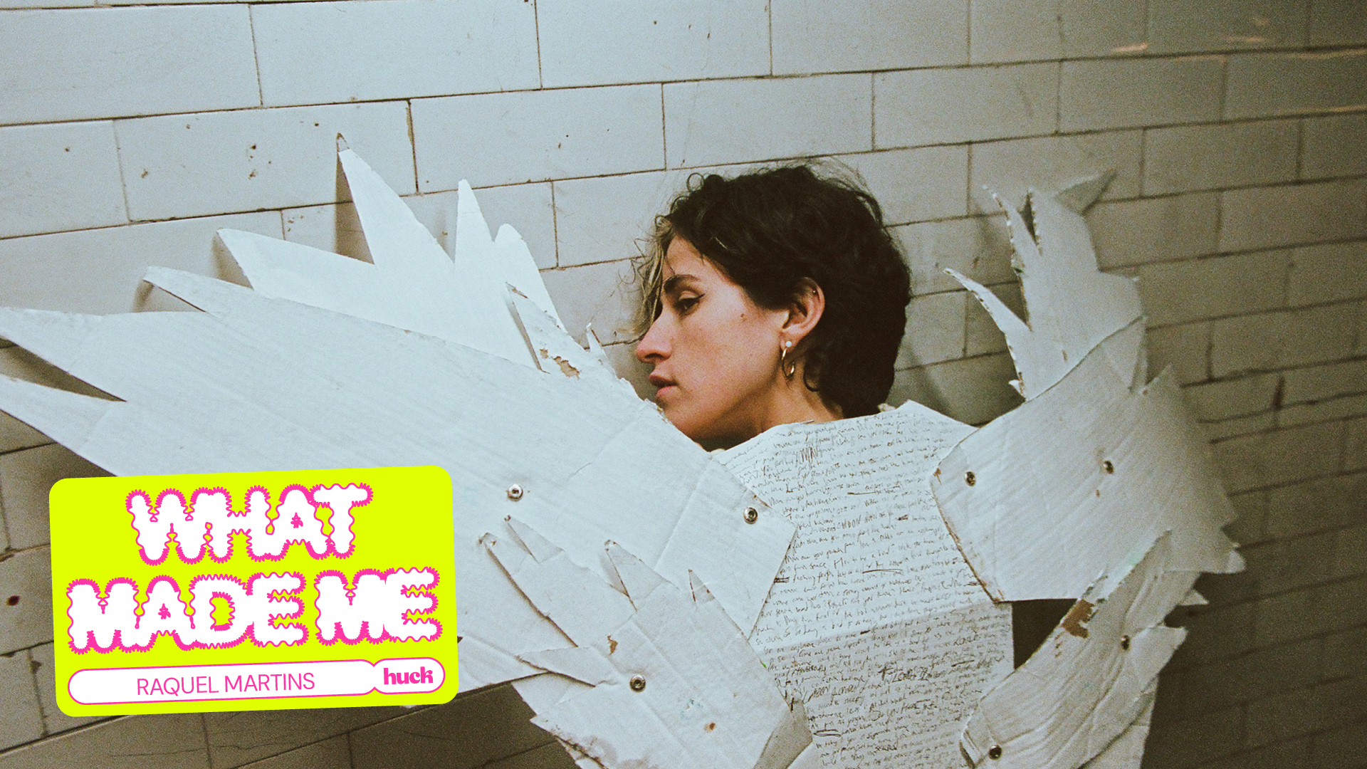Woman with short brown hair wearing white jumper, large white paper wings attached to shoulders, against white brick wall.
