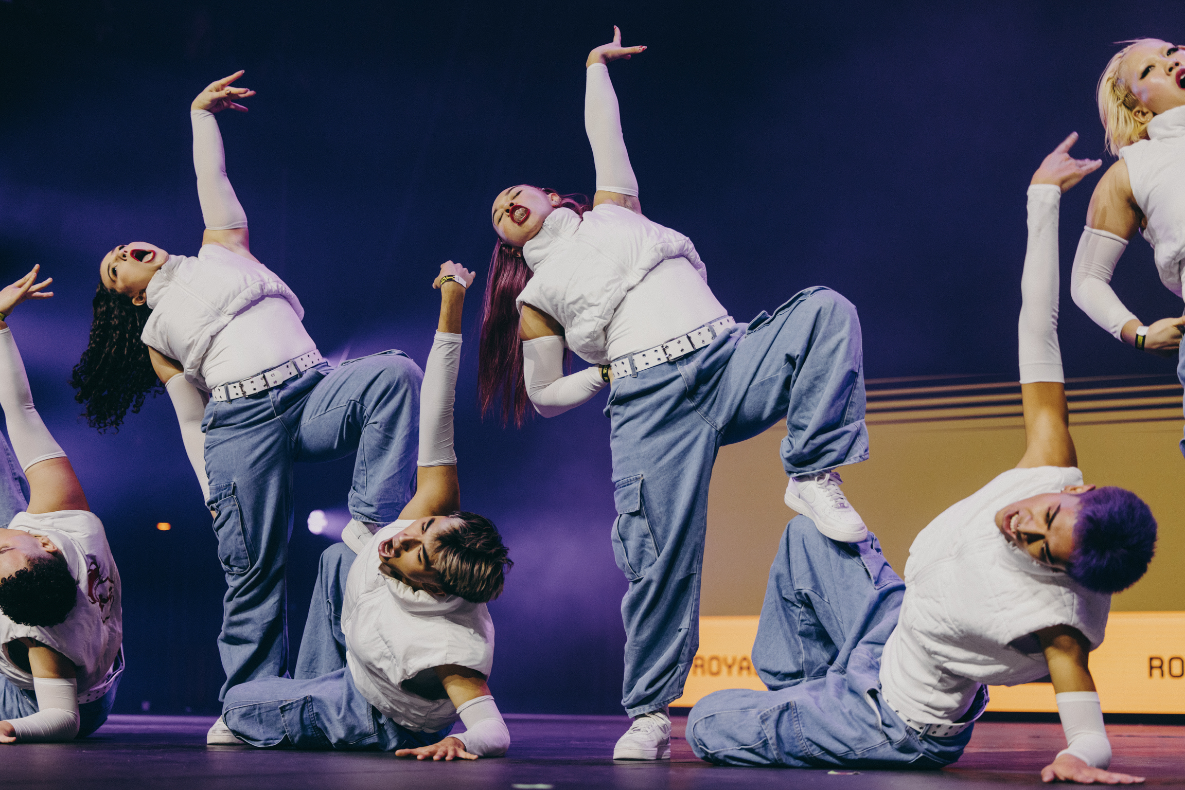 Group of dancers in white tops and grey trousers performing synchronised choreography on stage with purple lighting.