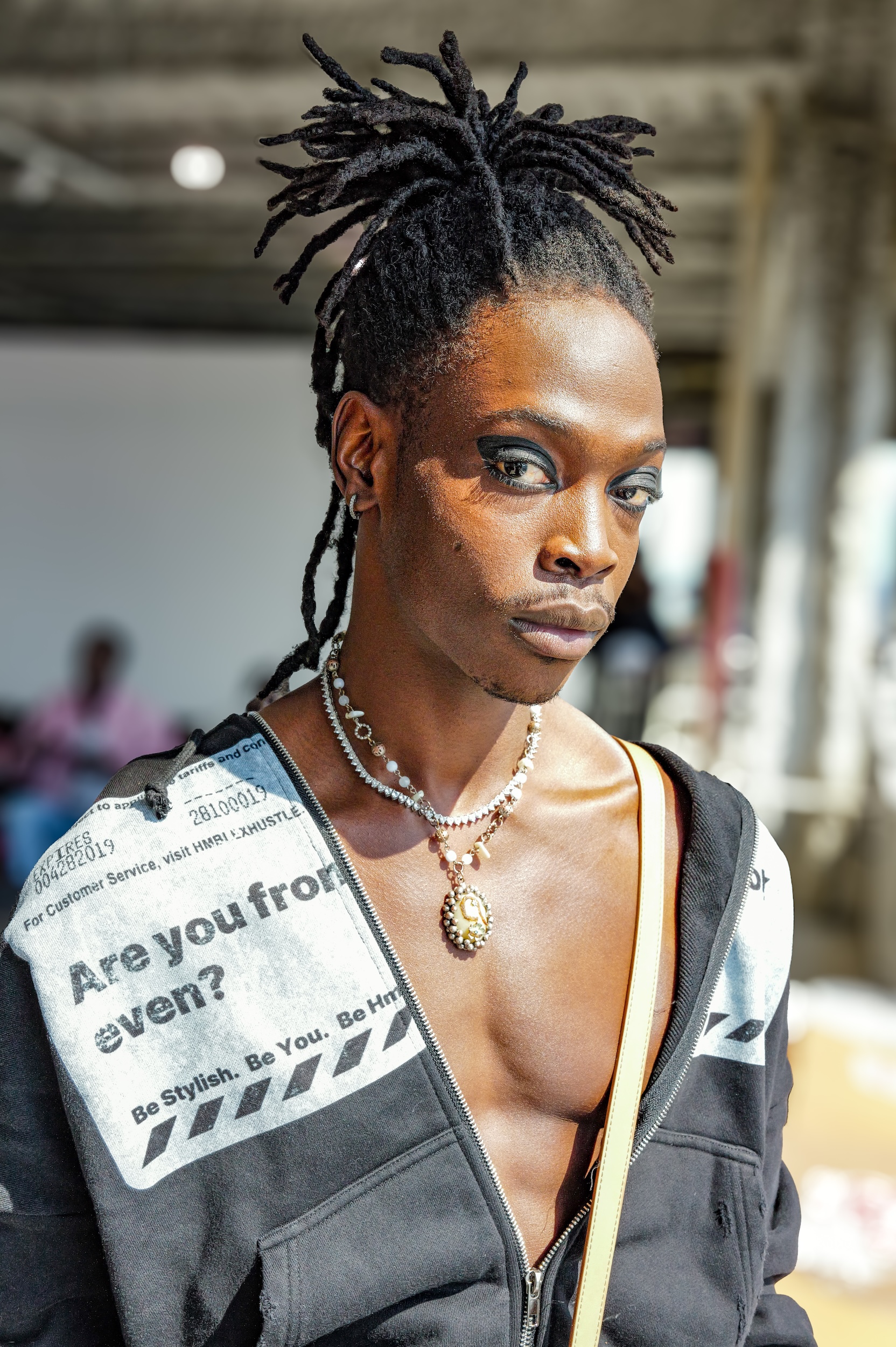 Black woman with locs styled upward wearing pearl necklaces and grey zip-up top with "Are you from even?" text visible on chest.