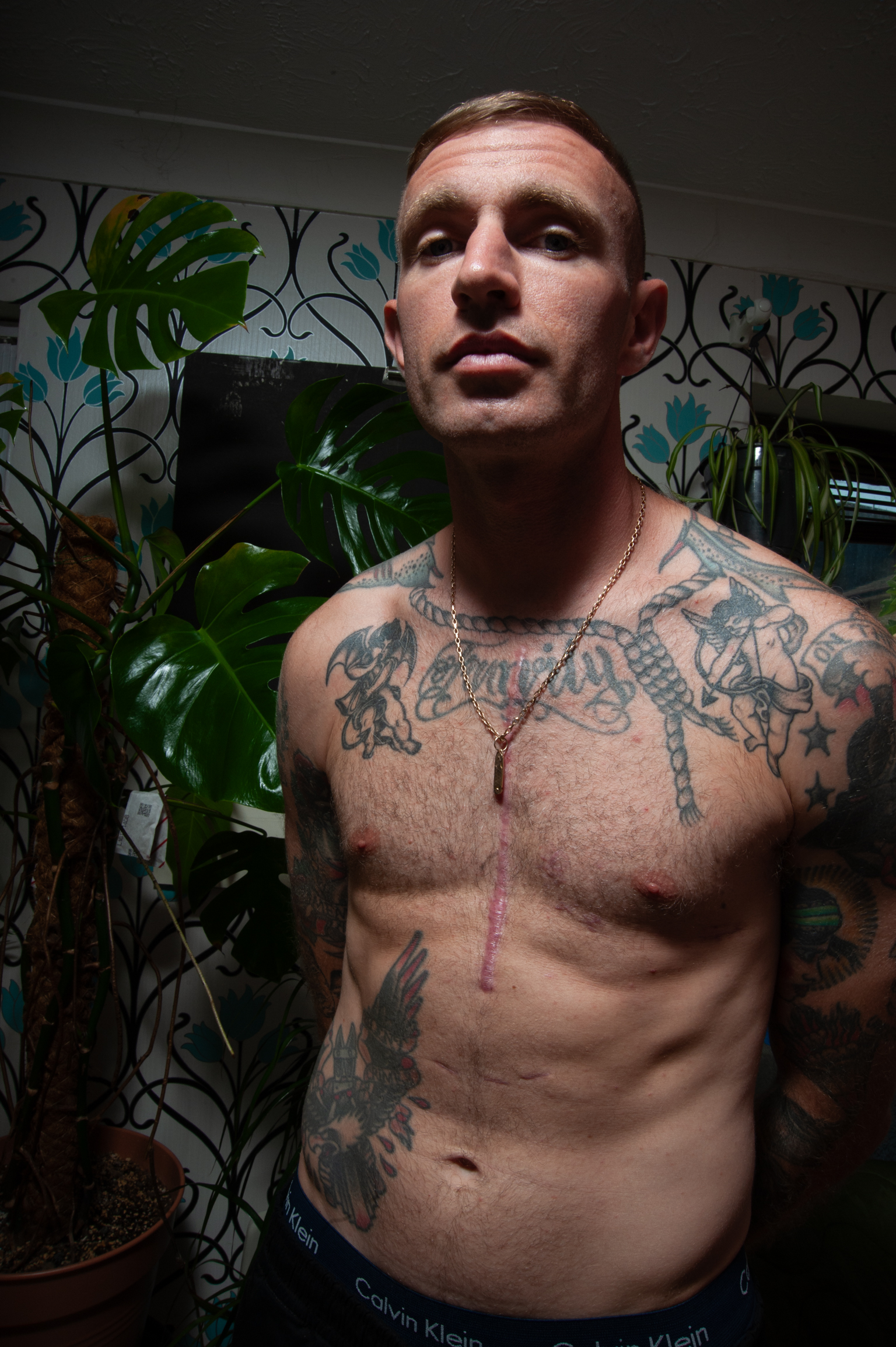 Shirtless tattooed man with chain necklace stands amongst green houseplants against floral patterned wallpaper.