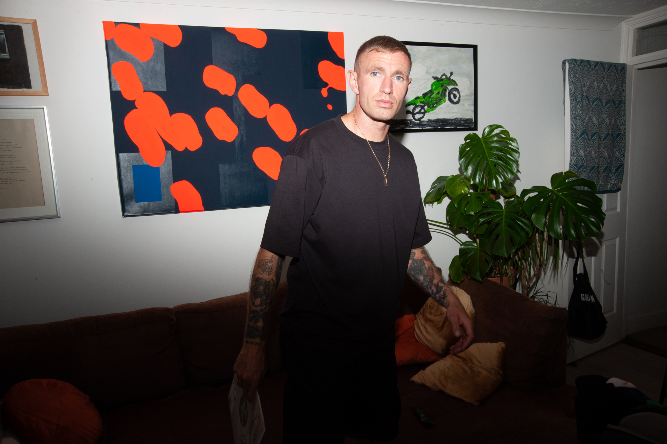 Man in black top standing in room with orange and blue abstract painting on wall, houseplants, and framed artwork.