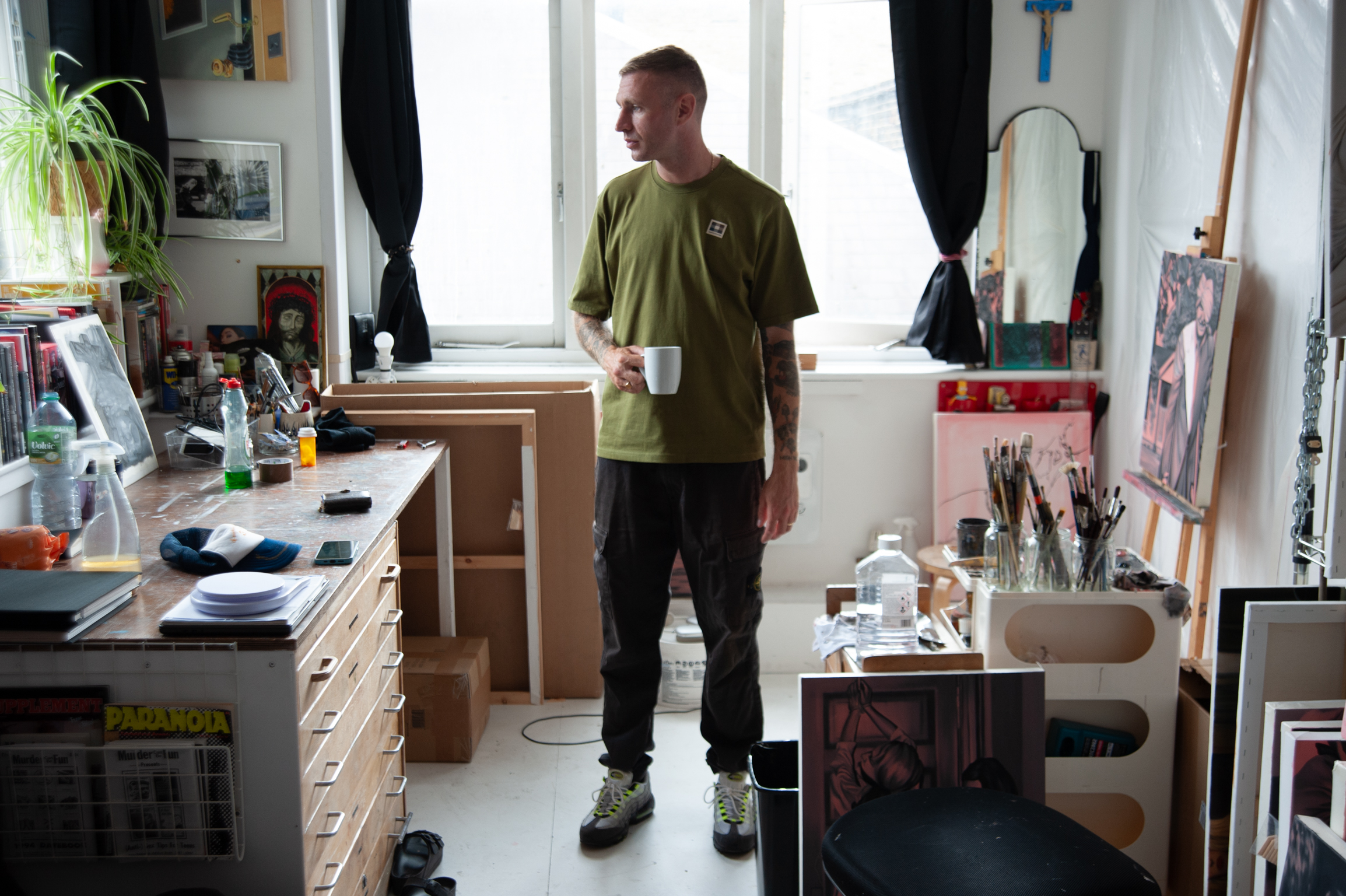 Man in olive green shirt holding white mug stands in cluttered art studio with easel, desk, supplies, and window with black curtains.