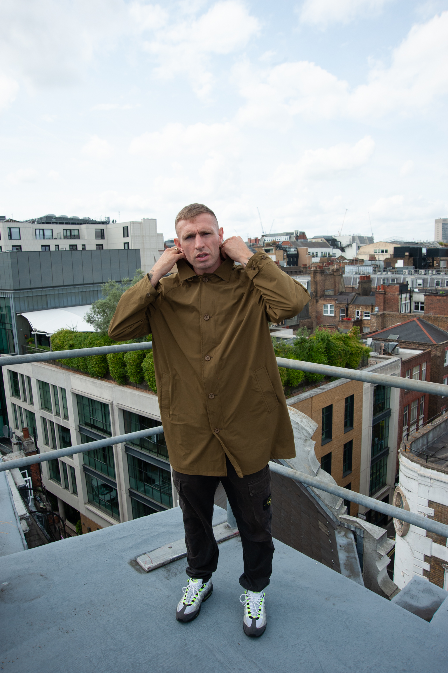 Man in olive green shirt adjusting collar on rooftop balcony overlooking mixed residential buildings under cloudy sky.