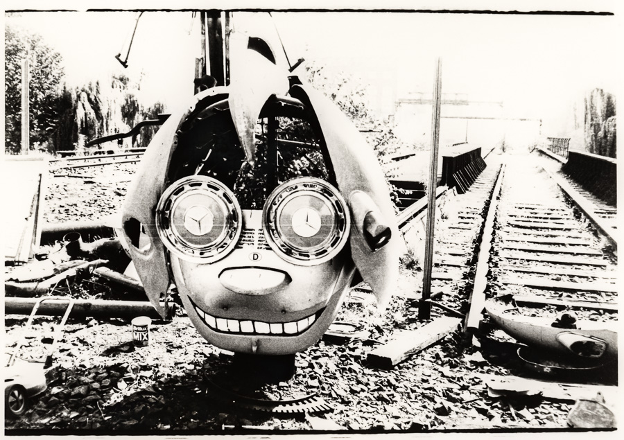 Black and white illustration of cartoon character head with large round goggles and grinning mouth on railway tracks with debris.