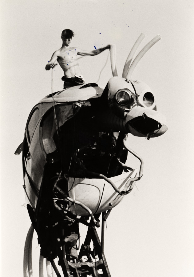 Black and white image: shirtless man stands atop large mechanical device with circular lenses and protruding metal components.