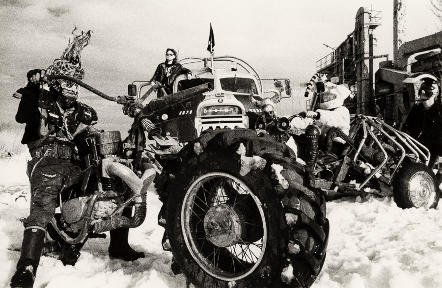 Black and white image showing people gathered around vintage motorcycles and vehicles in snowy conditions with overcast sky.