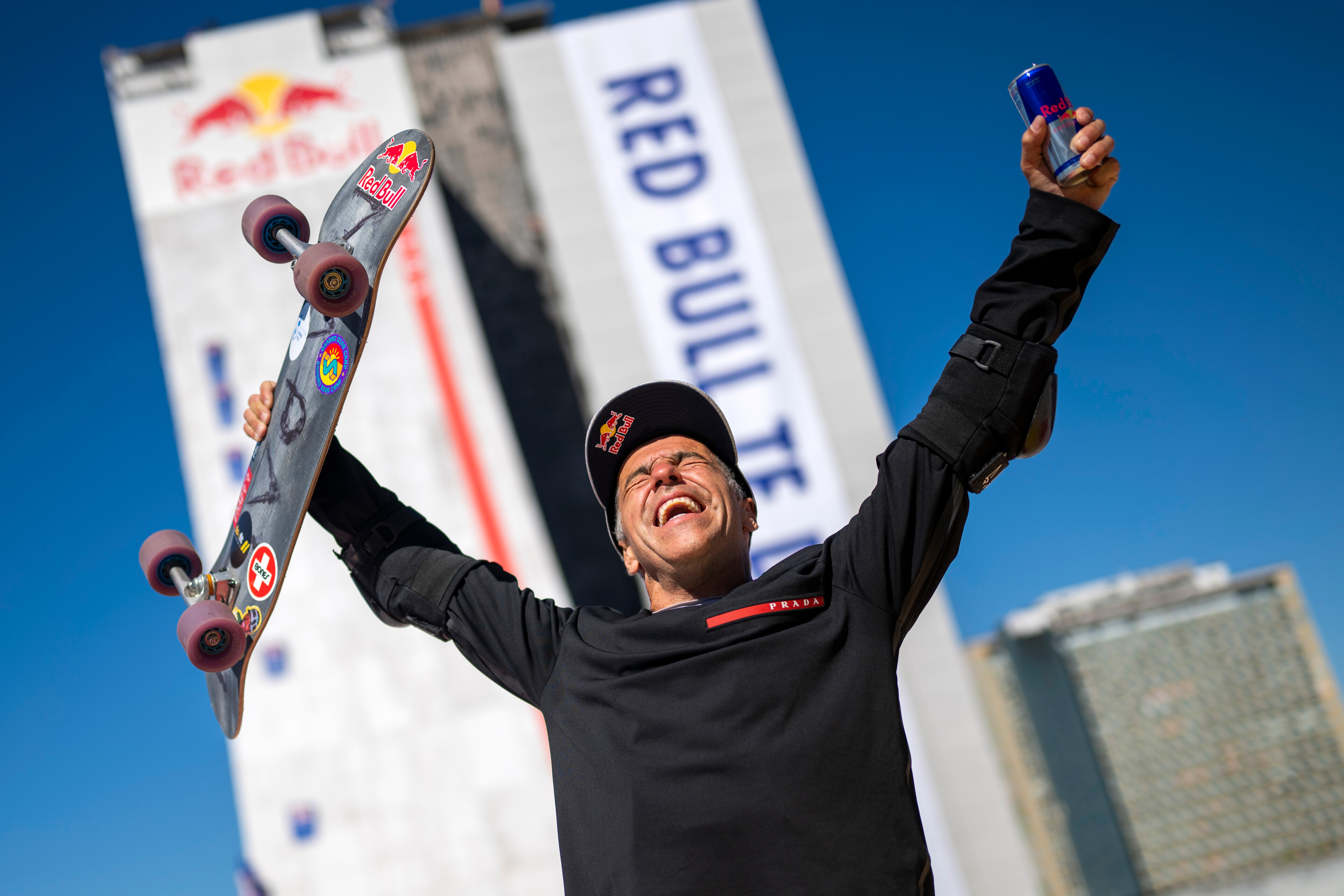 Man in black Red Bull uniform celebrating with arms raised, holding skateboard, against blue sky with Red Bull banner behind.