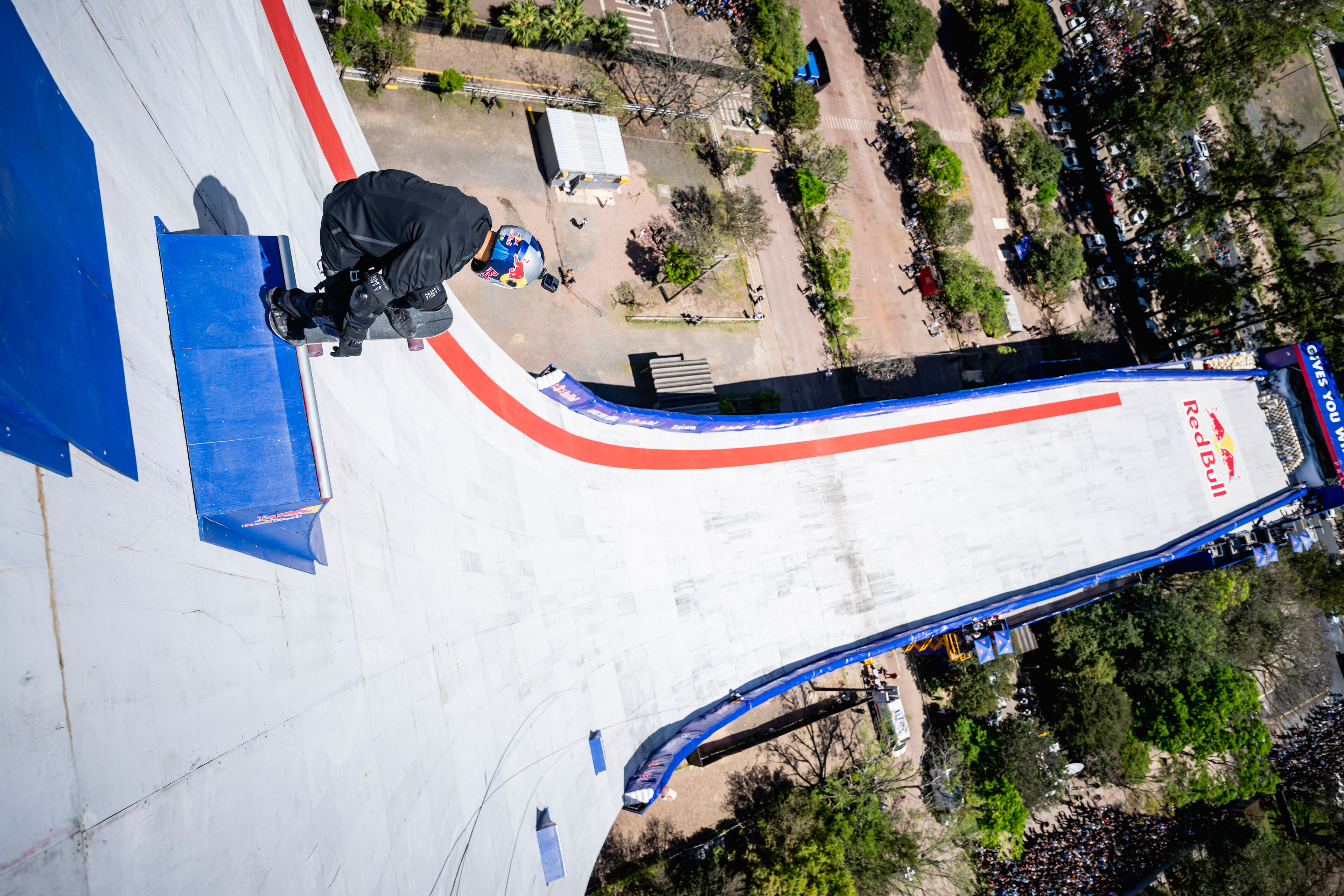 Aerial view of concrete skate park with blue, white and red curved surfaces, surrounded by trees and dirt paths.