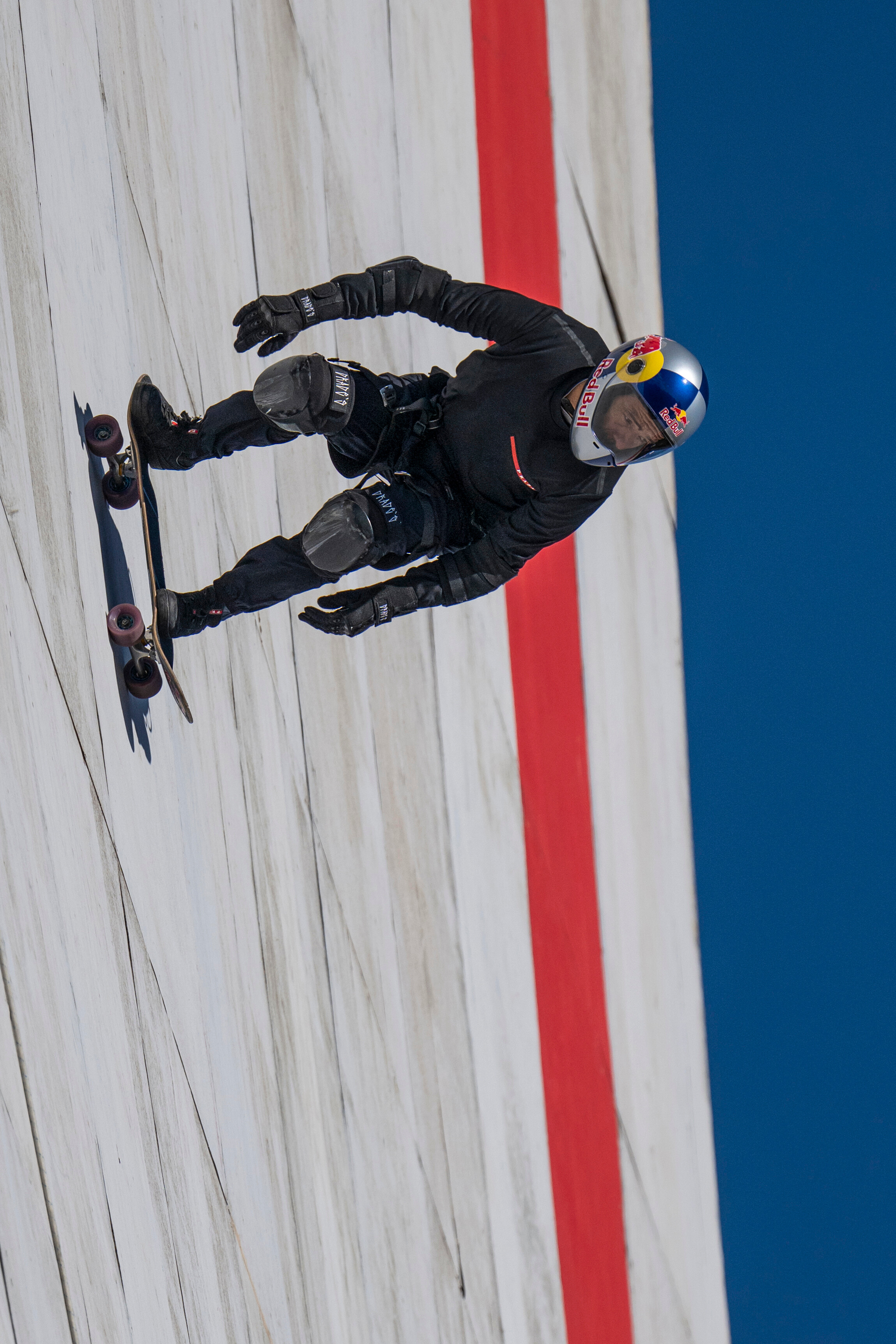 Skier in black kit with colourful helmet mid-jump on white slope with red boundary line and blue sky background.