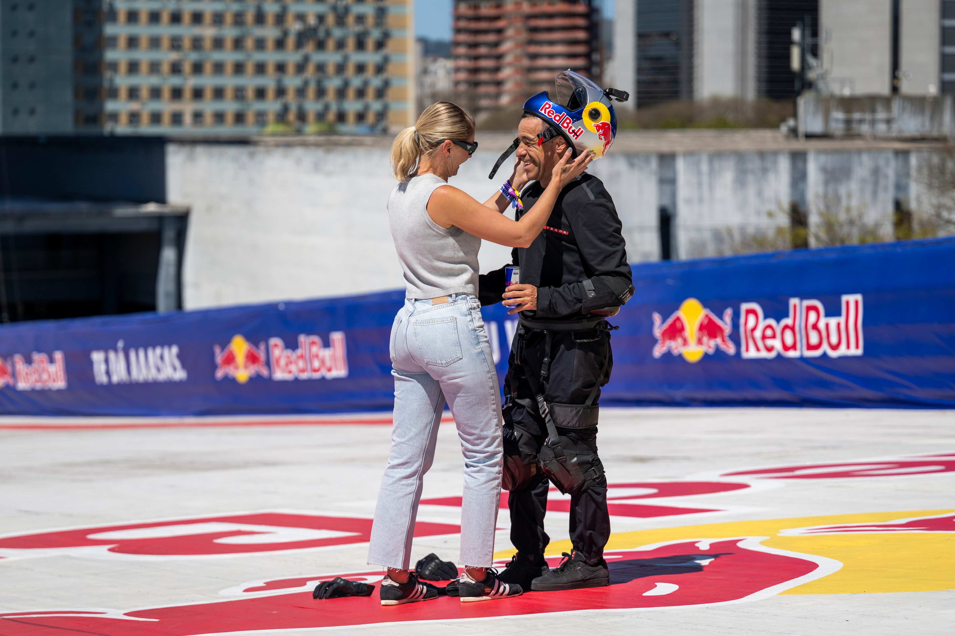 Two racing drivers embracing on red and white striped circuit. One wears white racing suit, other in black with colourful helmet.