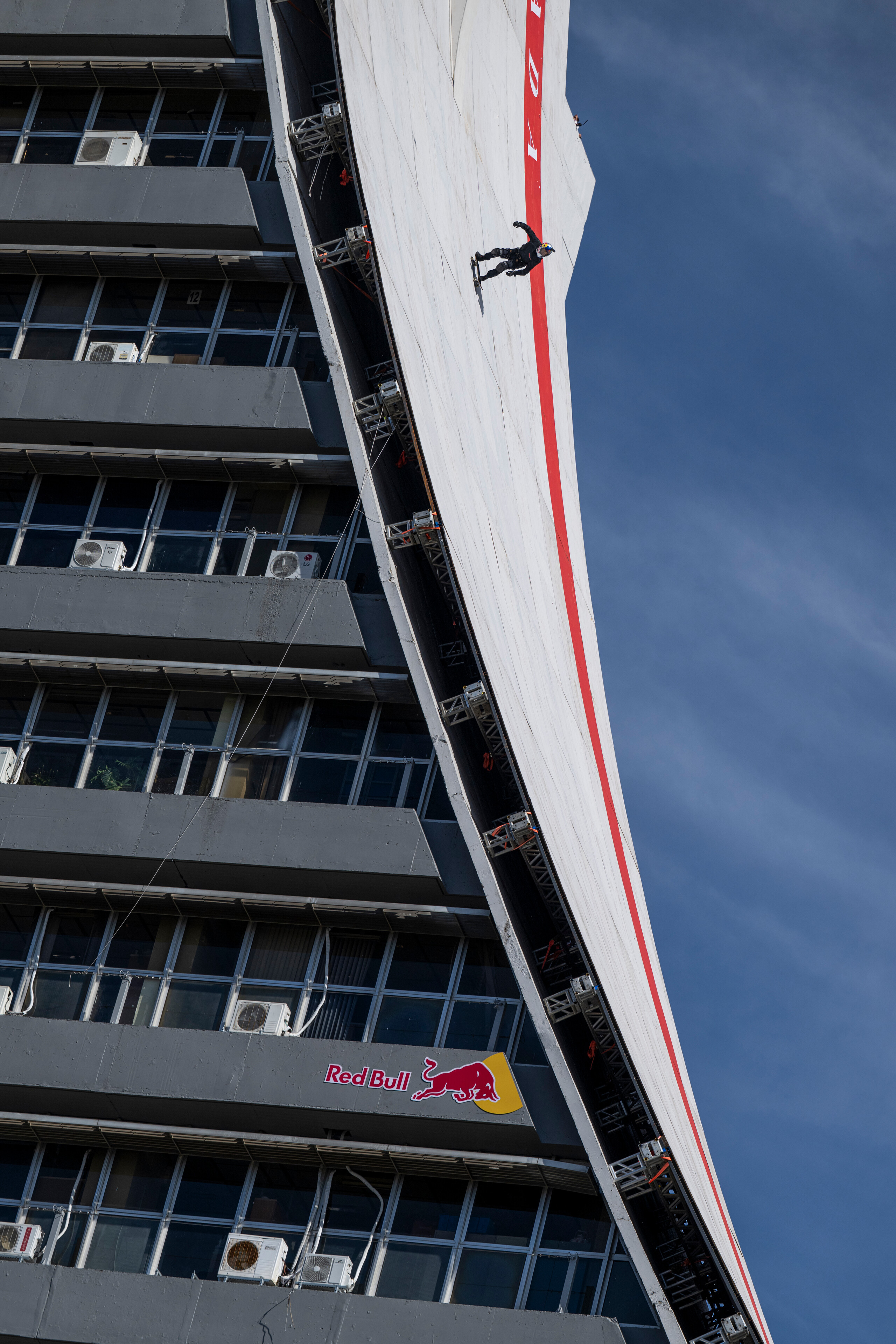 Person abseiling down white building wall with red stripe, Red Bull logo visible, blue sky background, shot from below.