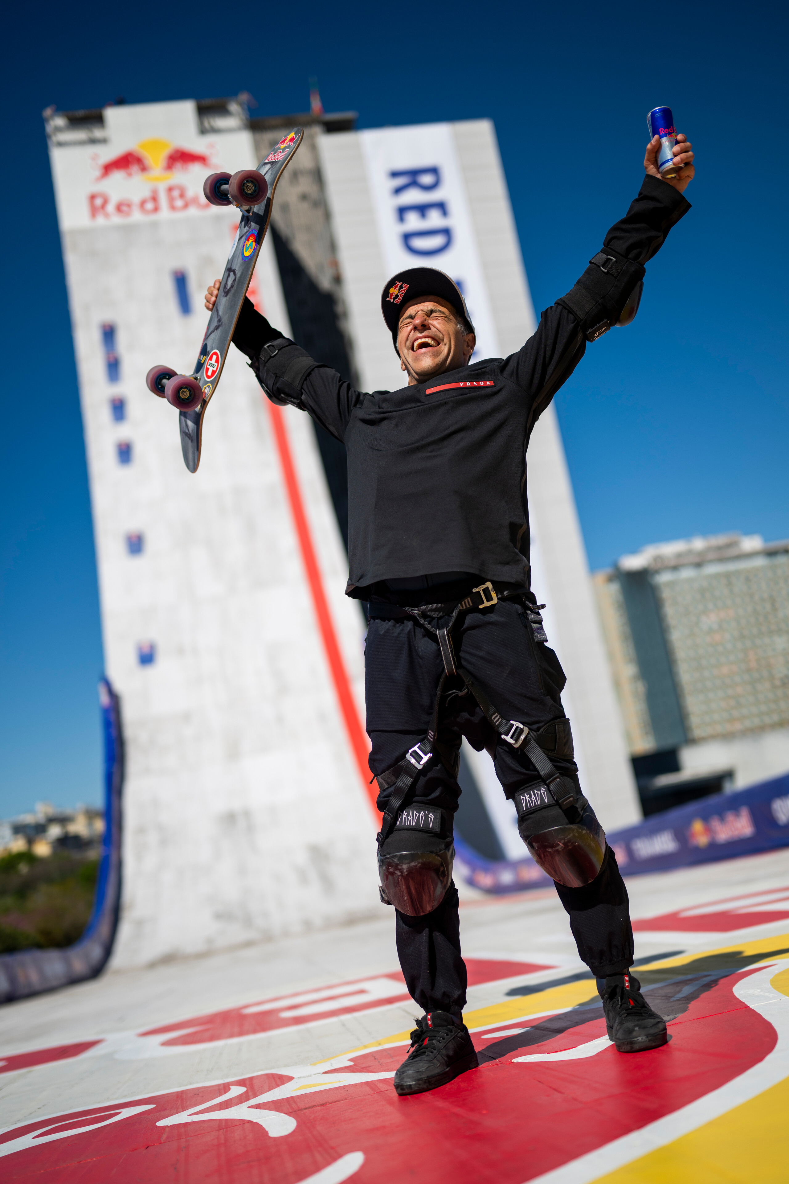 Skateboarder in black gear celebrates with arms raised, holding skateboard, standing on colourful platform with Red Bull ramp behind.
