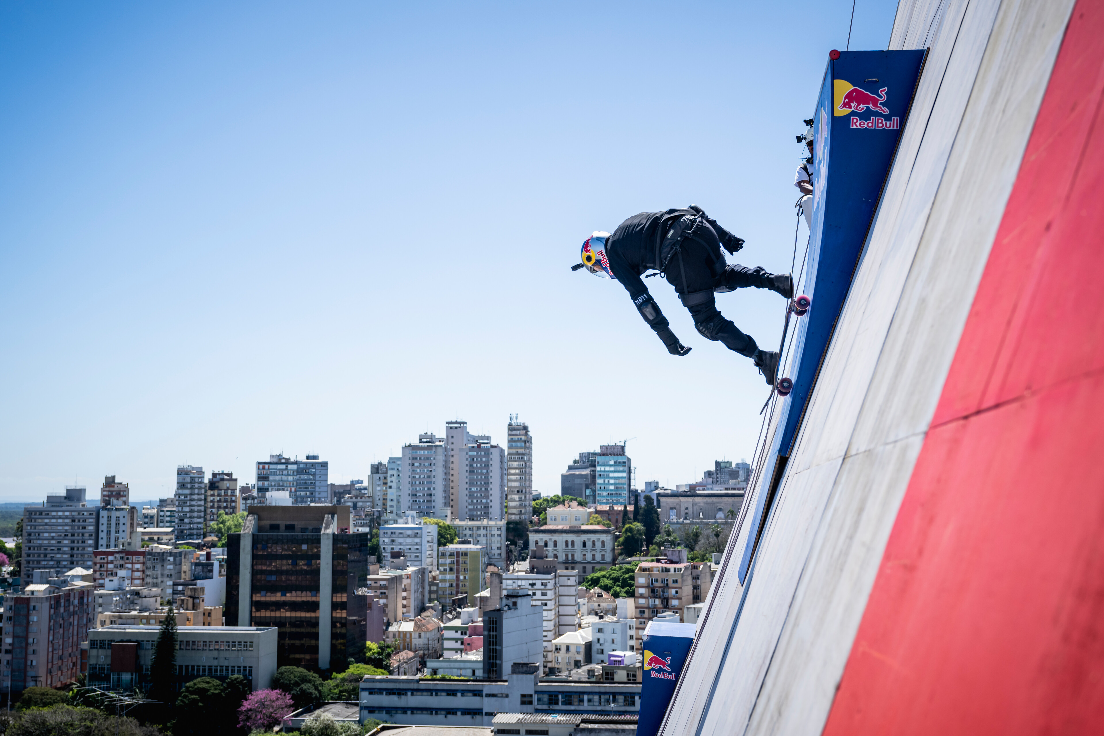 Person in black clothing rappelling down exterior wall of tall building with urban cityscape visible below against blue sky.