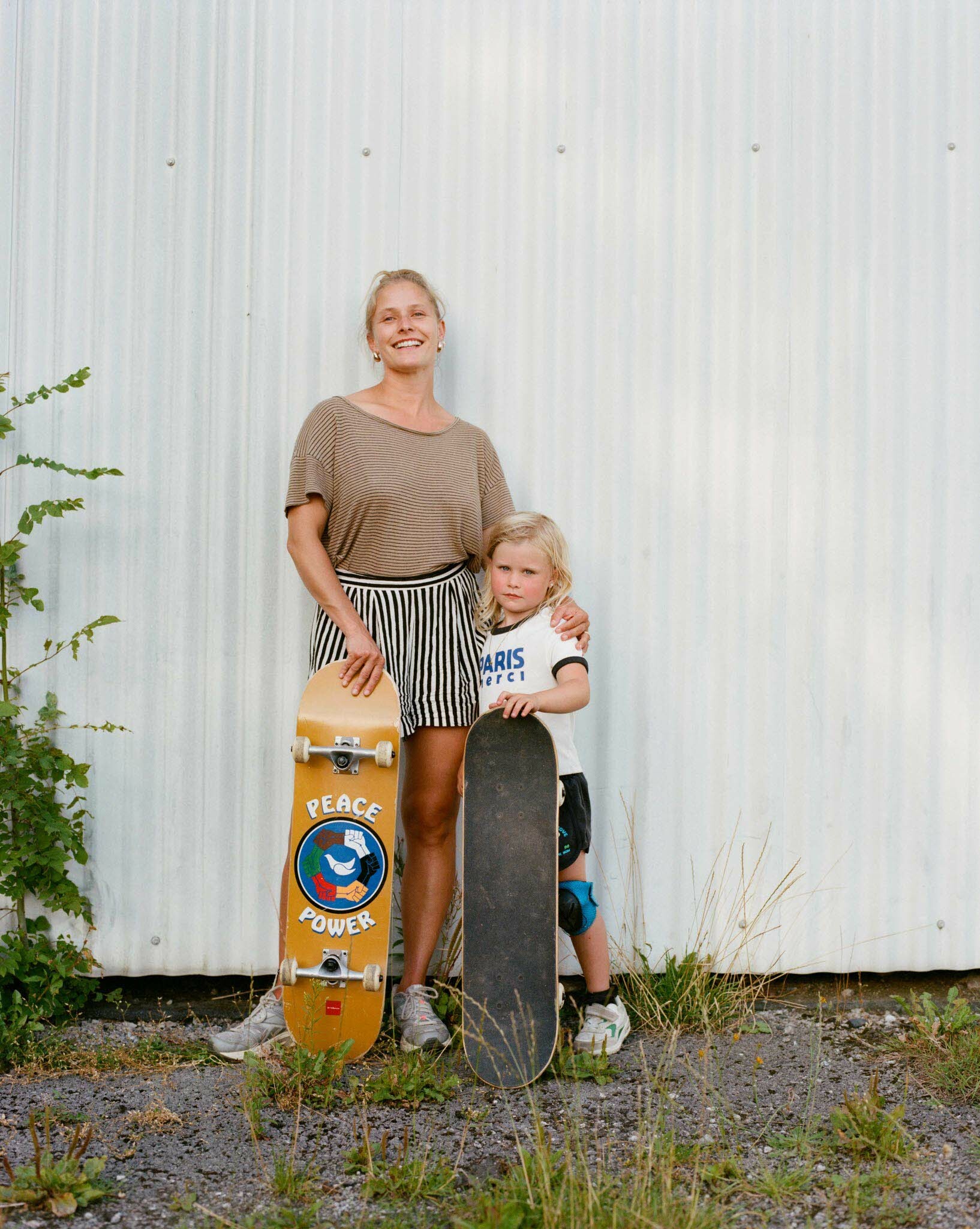 Woman in beige top and striped shorts standing with young child beside skate ramp with "Peace Power" sticker.
