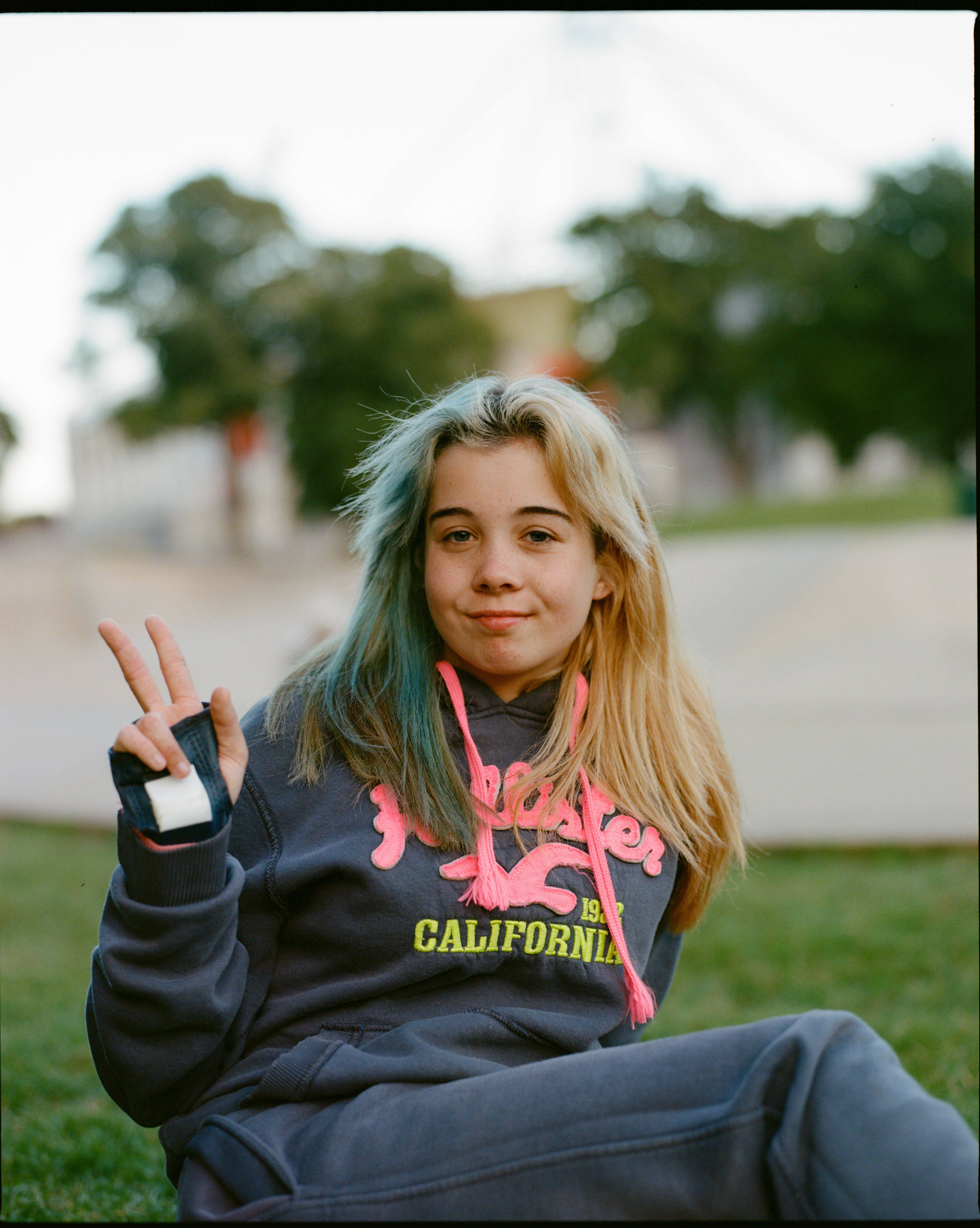 Smiling young female in California hooded top making peace sign against blurred nature background.