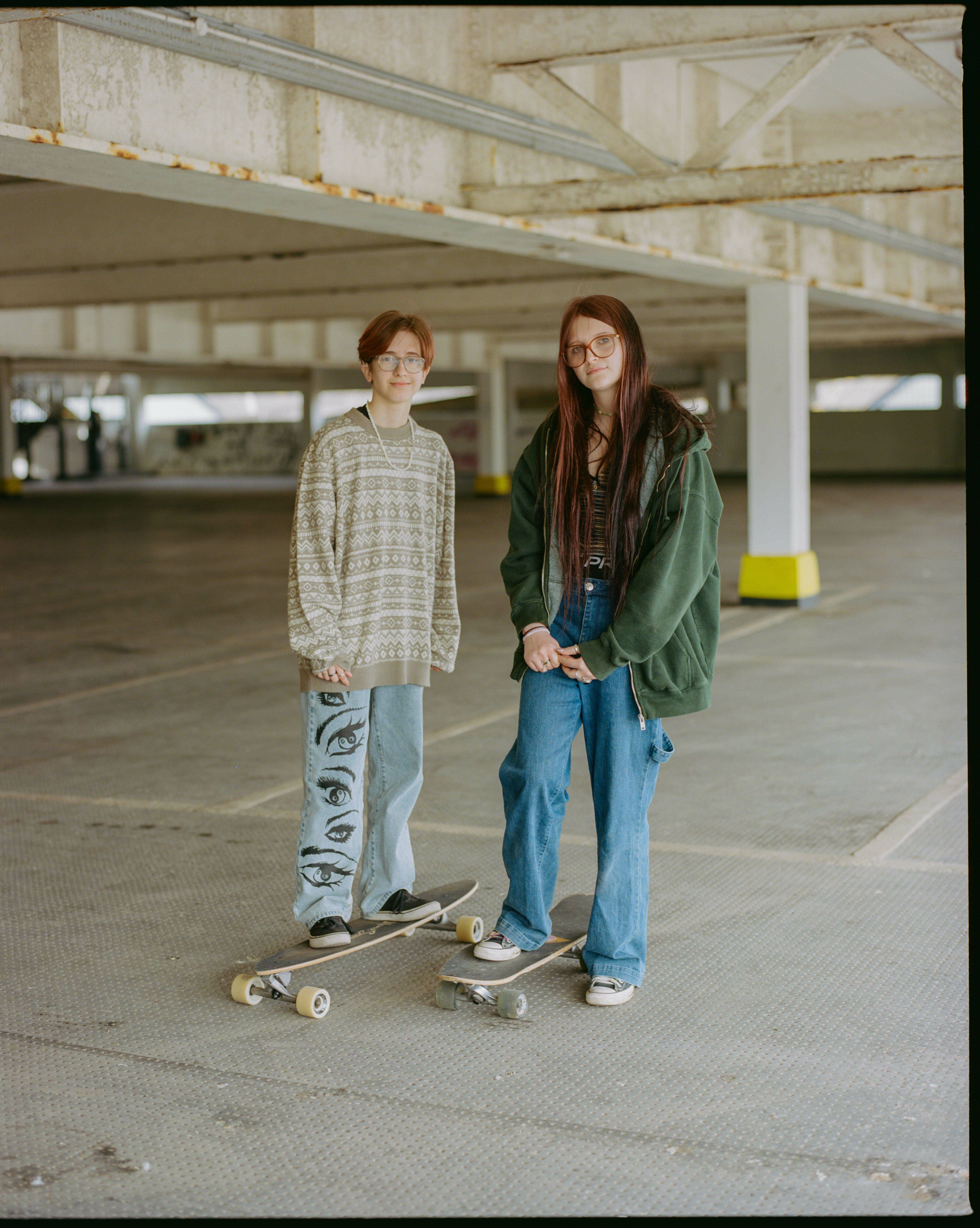 Two young women, one wearing a patterned jumper and the other a green coat, standing together with skateboards in a car park.