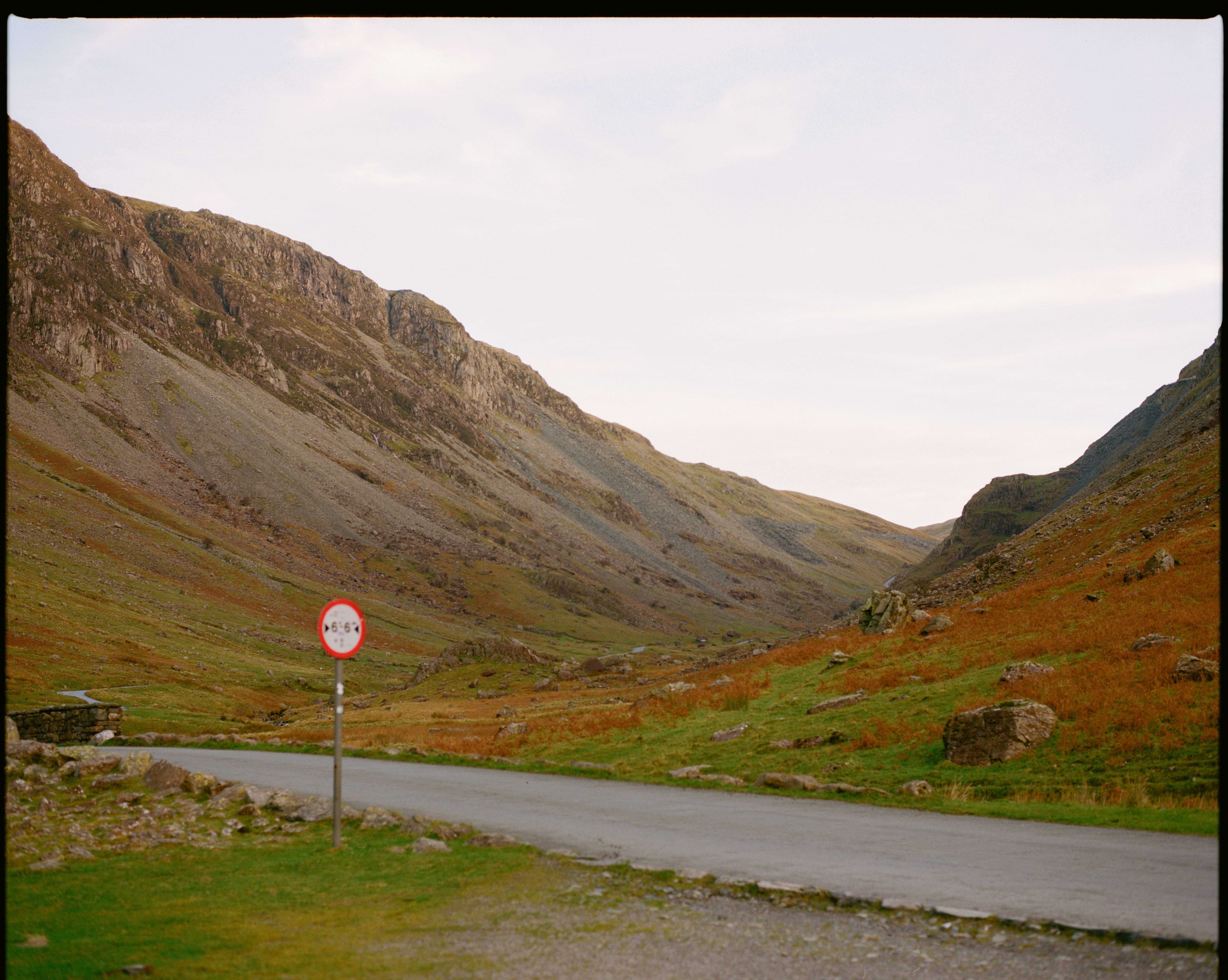 Winding road through hilly, grassy countryside with rocky cliffs in the background.