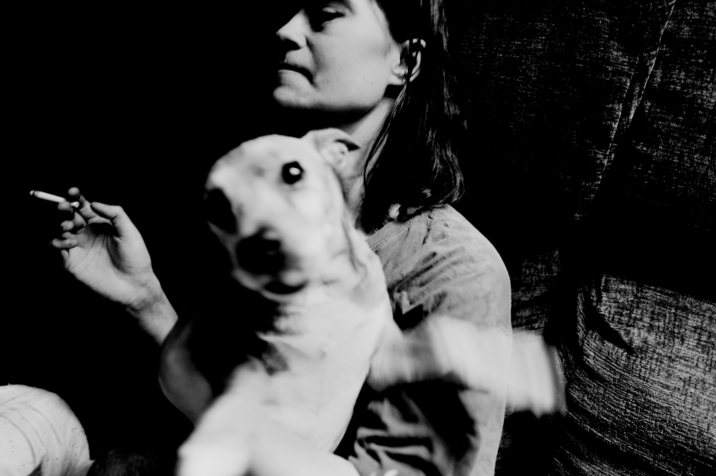 Black and white image of woman holding cigarette with white dog's head prominently featured in foreground, creating dramatic contrast.