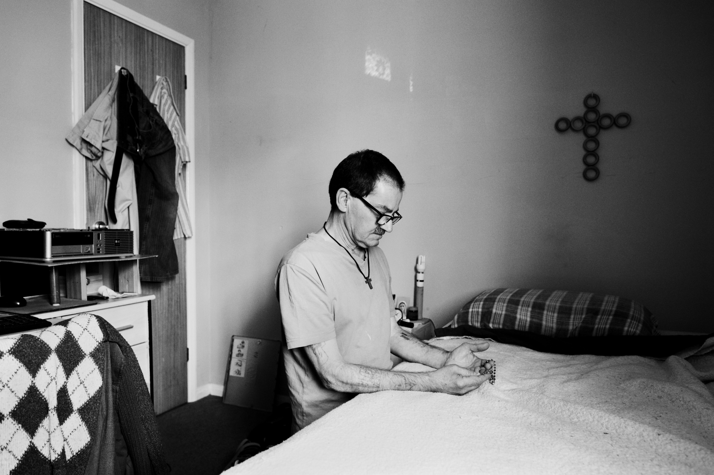 Man in glasses and light jumper sits on bed reading in sparse room with cross on wall, clothes hanging in doorway.