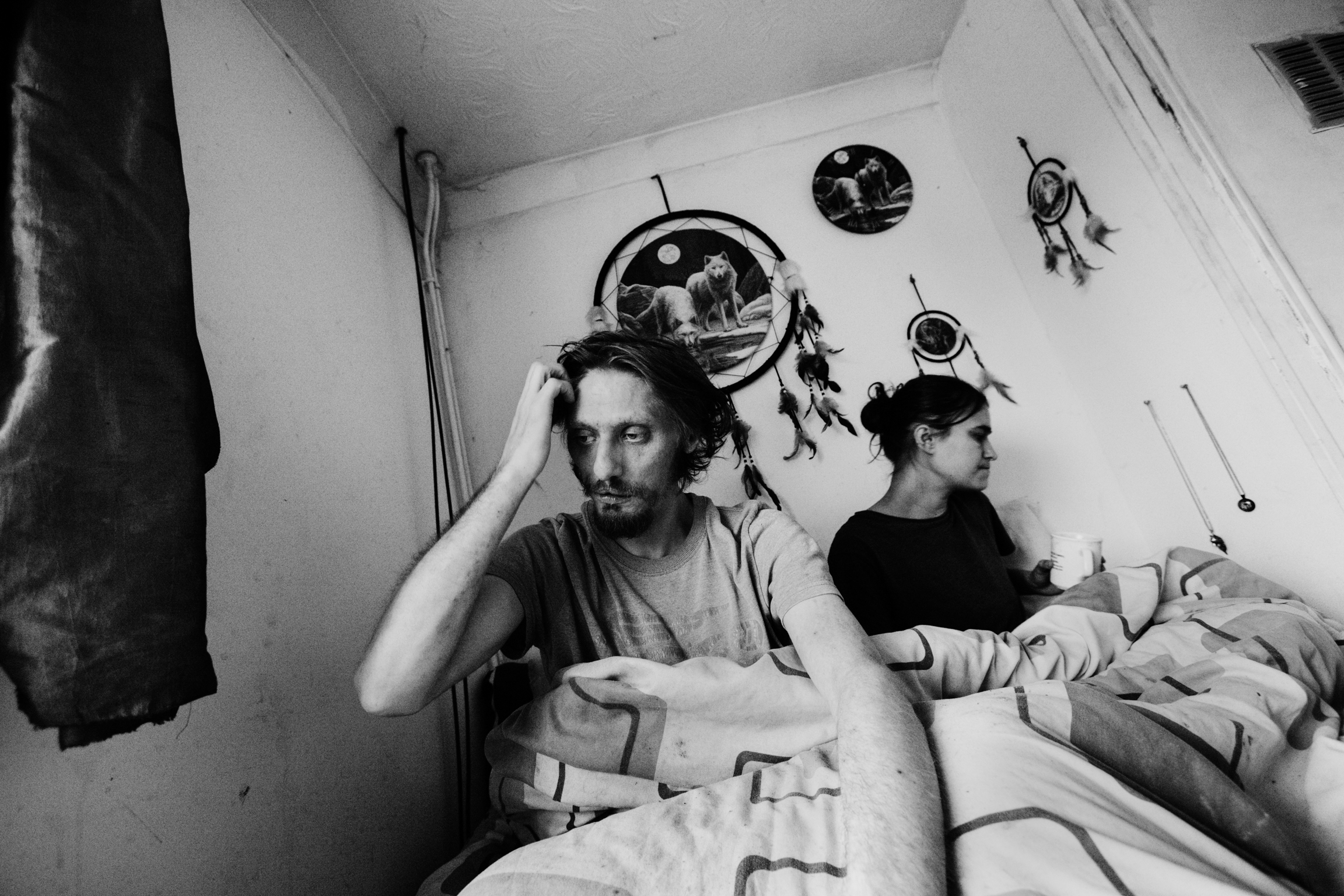 Black and white image of two people in bed. Man on left holds head in hand, woman on right lies down. Circular wall decorations above headboard.