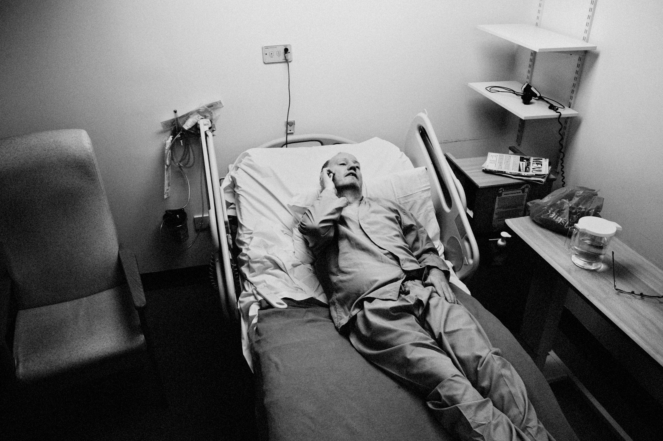 Black and white hospital room with patient in bed, chair on left, medical equipment and bedside table with items on right.