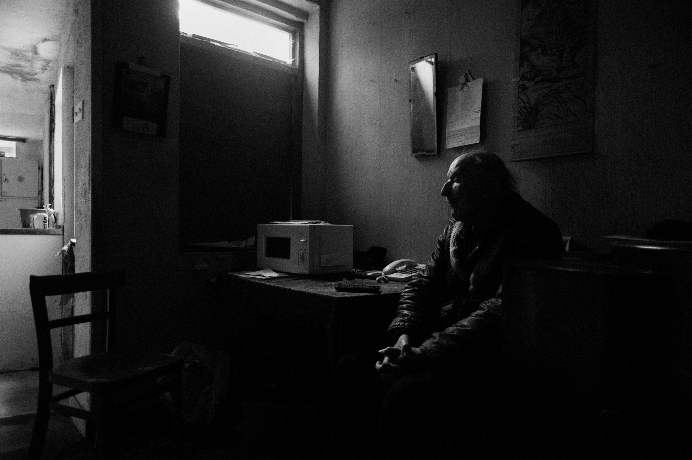 Person silhouetted at desk in dimly lit room with bright window, wooden chair visible, black and white photograph.