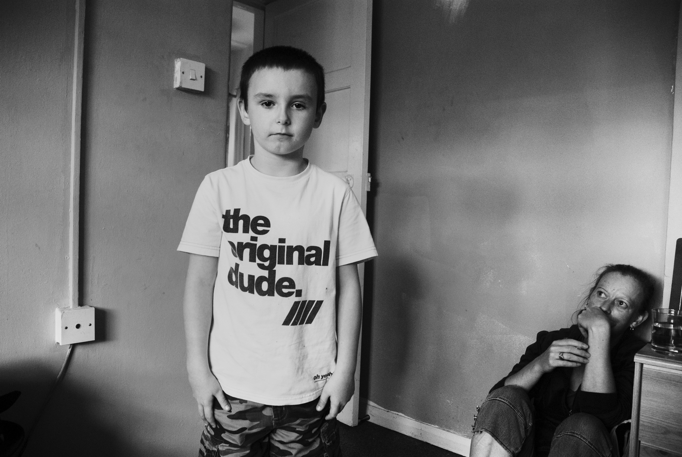 Young boy in "the original dude" t-shirt standing in room, man with drink seated at table in background, black and white image.
