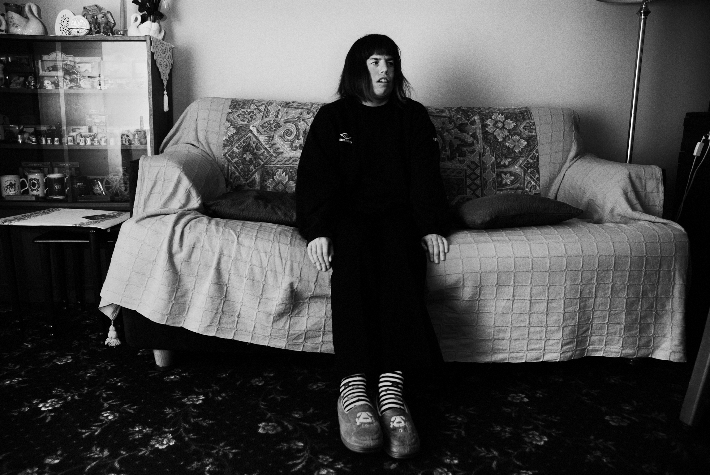 Person with long dark hair in black clothing sits on sofa with floral pattern, striped socks visible. Black and white image.
