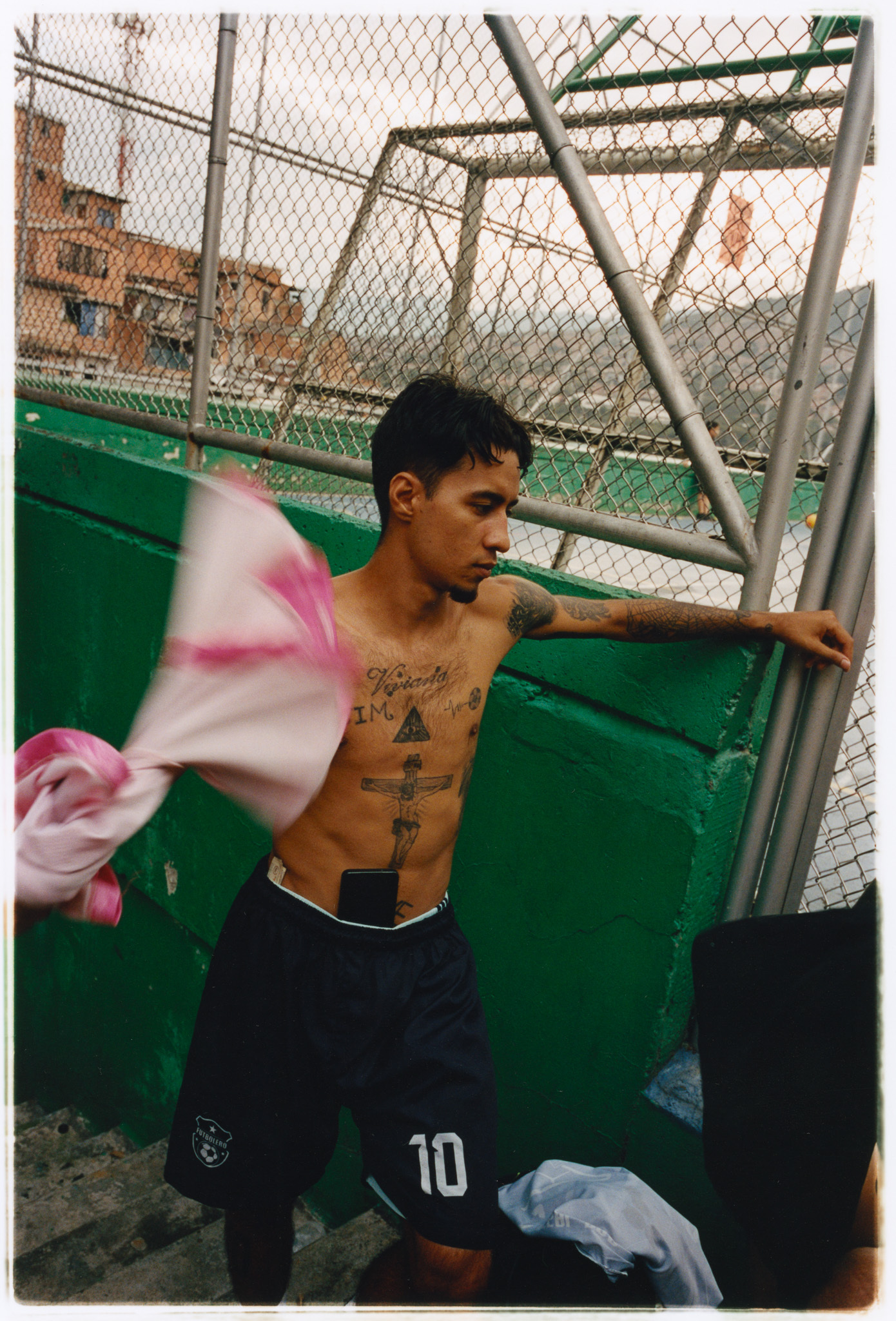 Man standing in front of a green fence, shirtless and with visible tattoos.