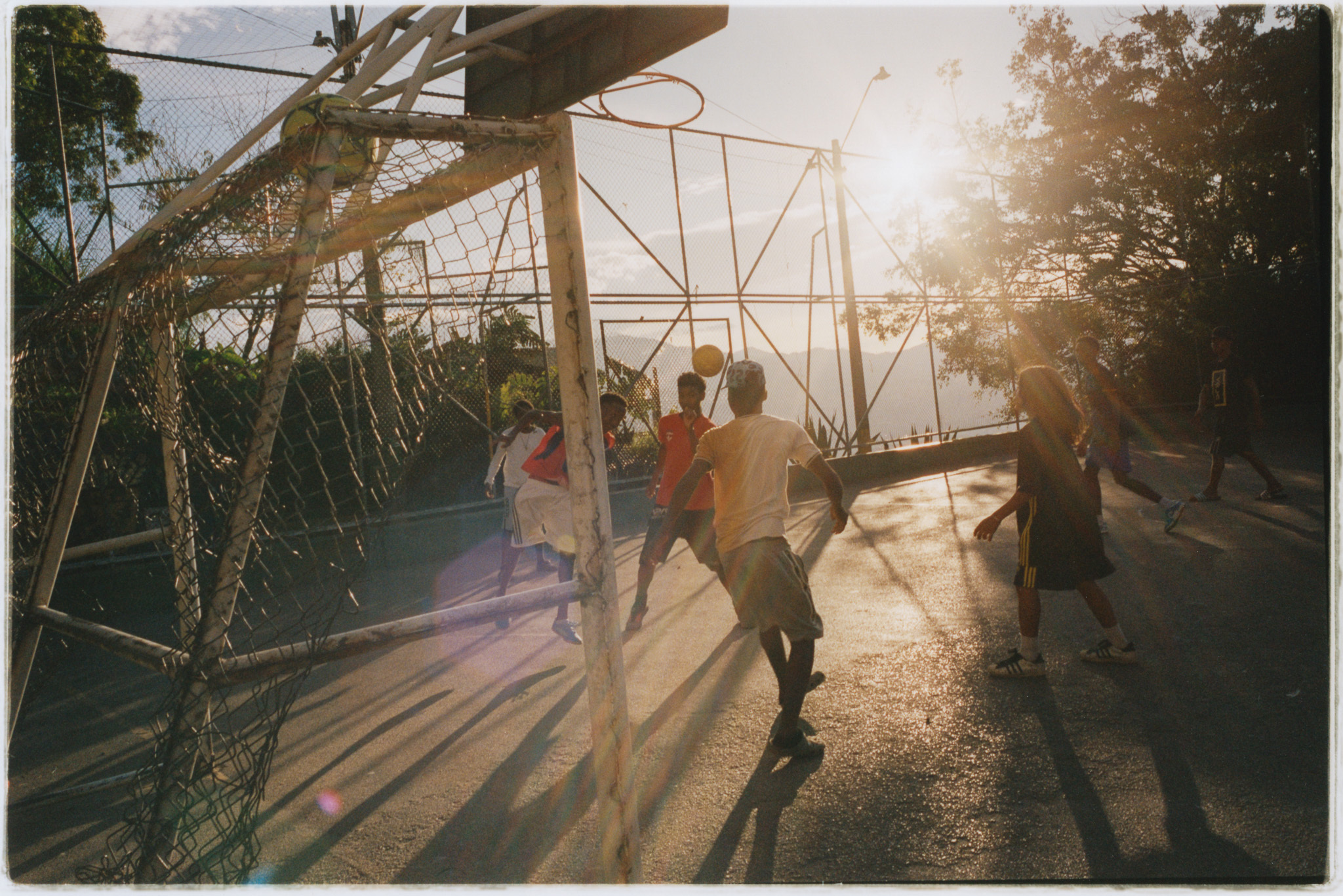 Rustic footbridge across a river, with people crossing it on a sunny day. Warm tones and shadows suggest an outdoor, natural setting.