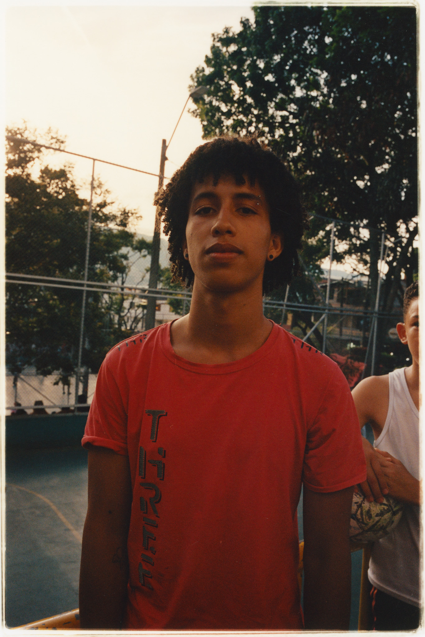 Young man wearing a red T-shirt with text, standing in an outdoor setting with trees in the background.