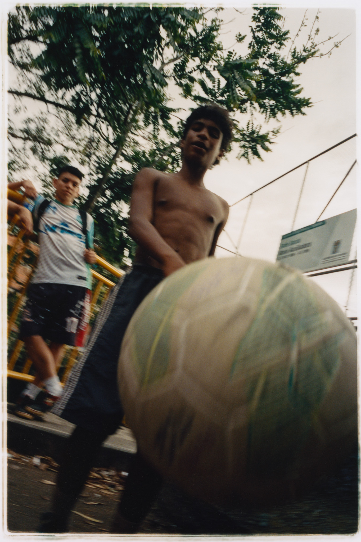 Two shirtless young men, one in the foreground holding a large inflatable ball, with a wooded area in the background.