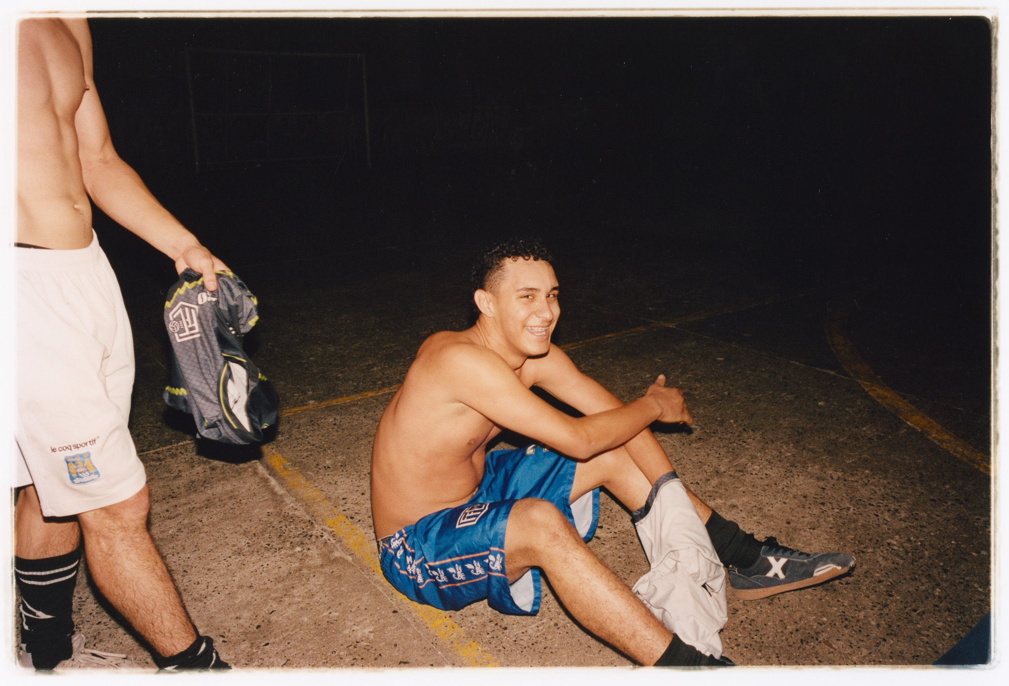 A shirtless man sitting on the ground, wearing blue shorts and socks, in a dark outdoor setting.