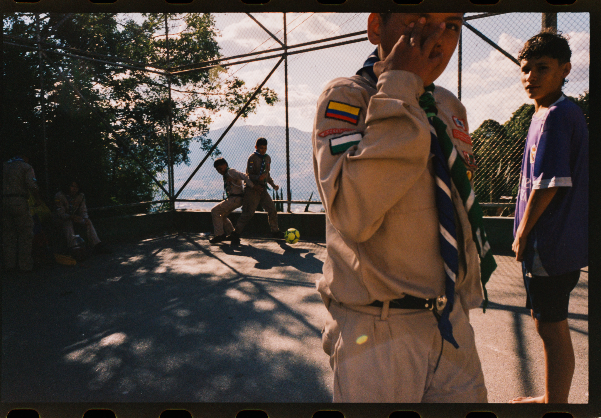 A group of people outdoors in casual clothing, with trees and shadows visible in the background.