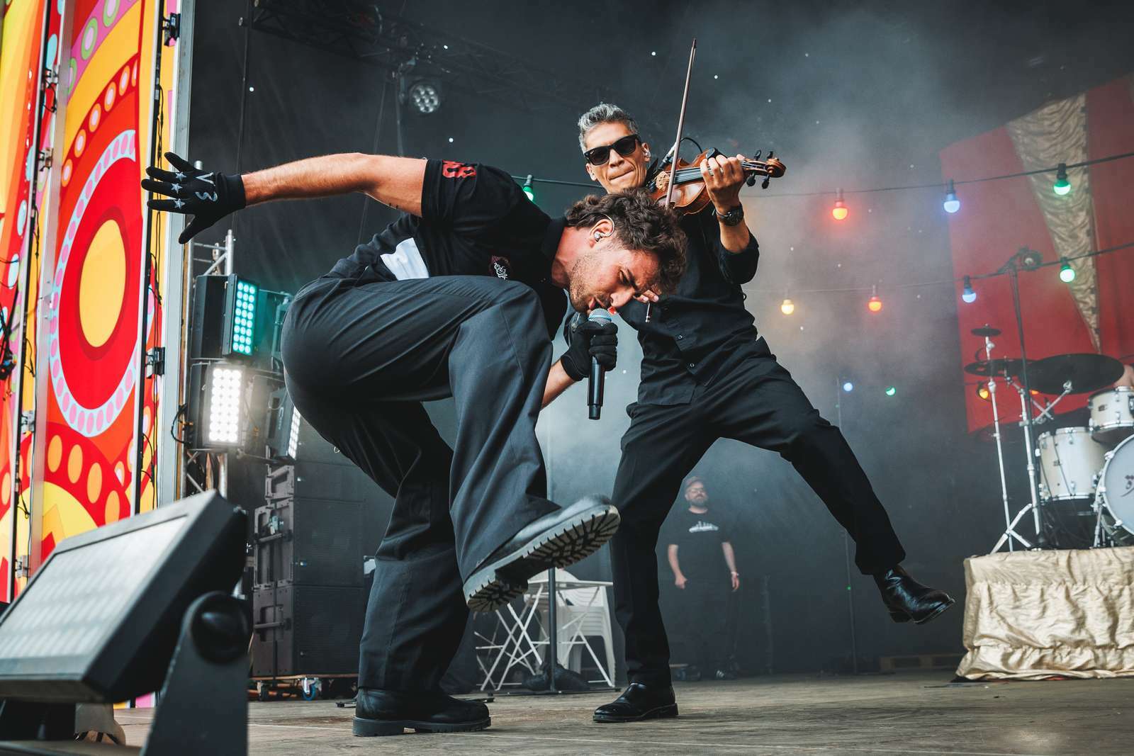 Two musicians performing on stage - one playing violin whilst standing on the other's back. Colourful backdrop with red and yellow patterns.
