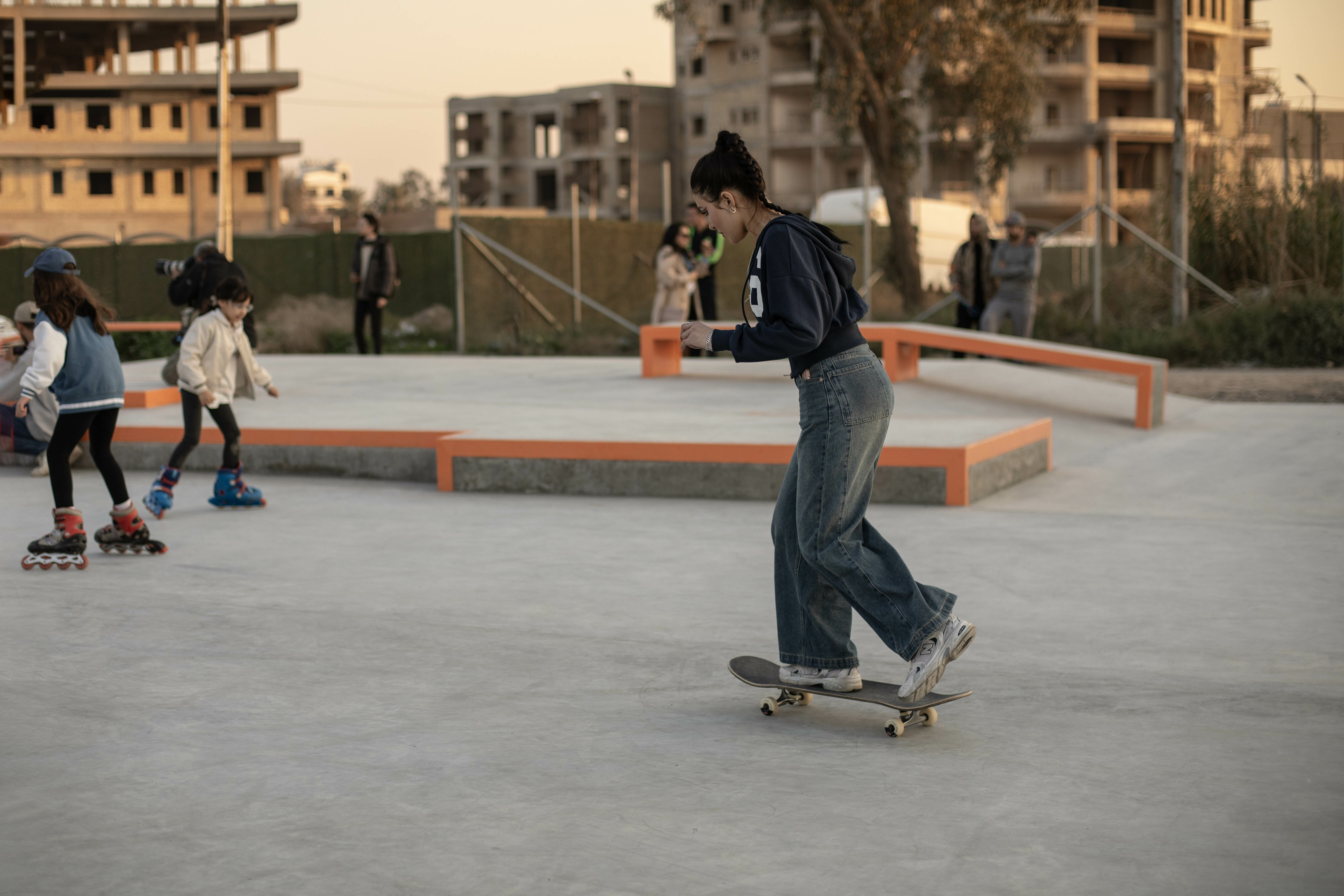 Skaters at skate park, some on rollerblades and one on a skateboard, amidst concrete structures and buildings in background.