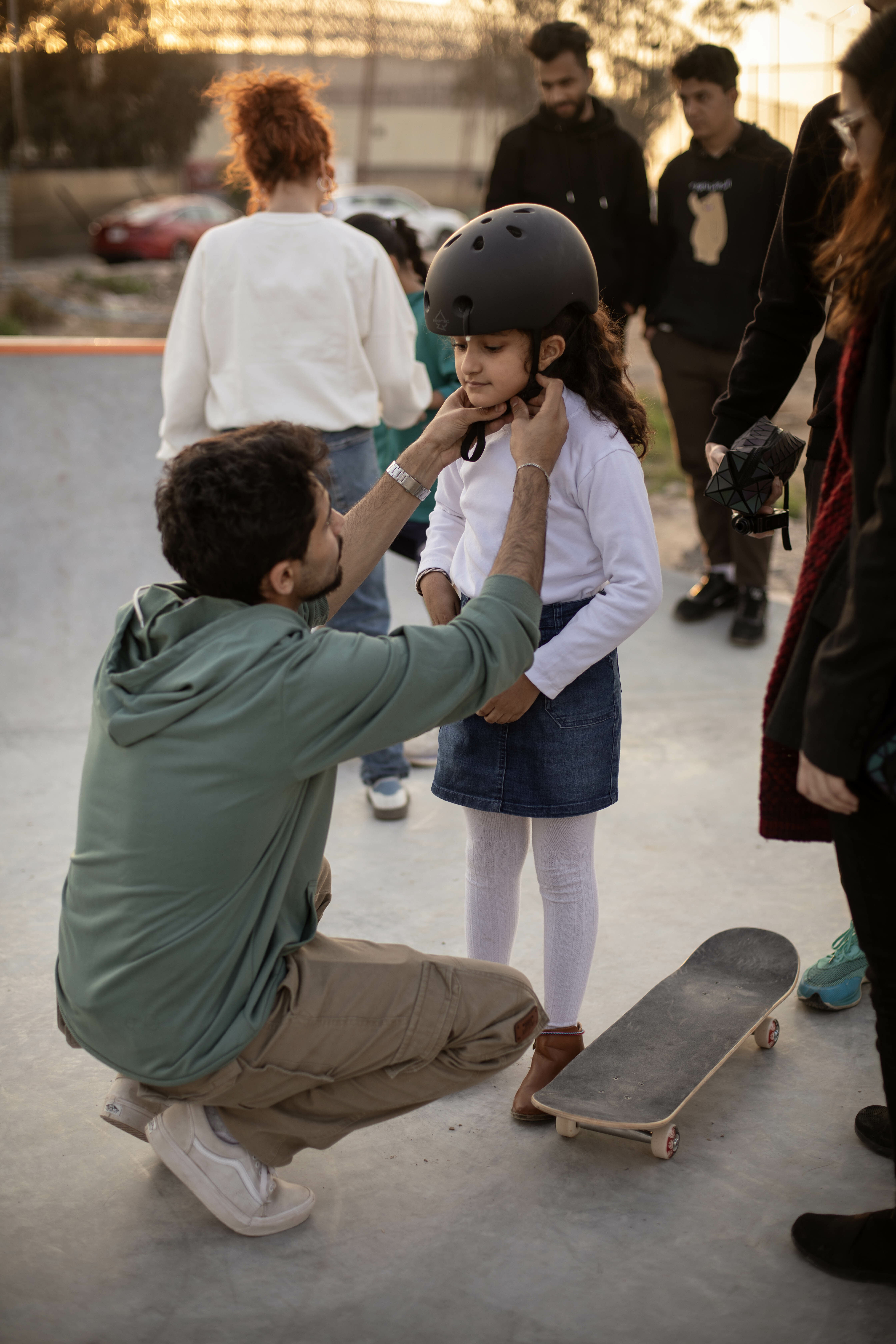 A man helping a girl put on a helmet on a skatepark with other people in the background.