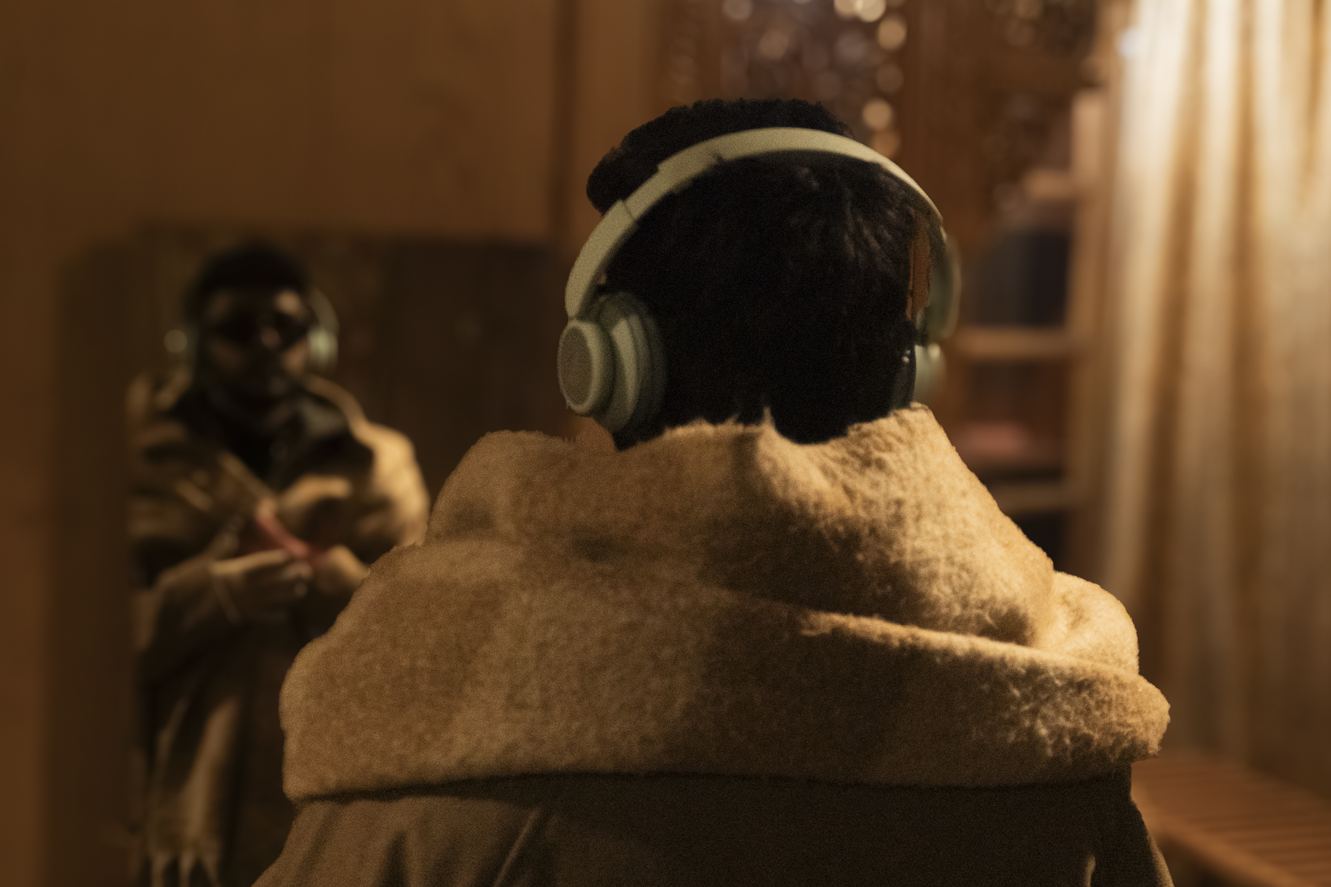 Person wearing headphones and beige hoodie sits with back to camera, another person visible blurred in background indoors.