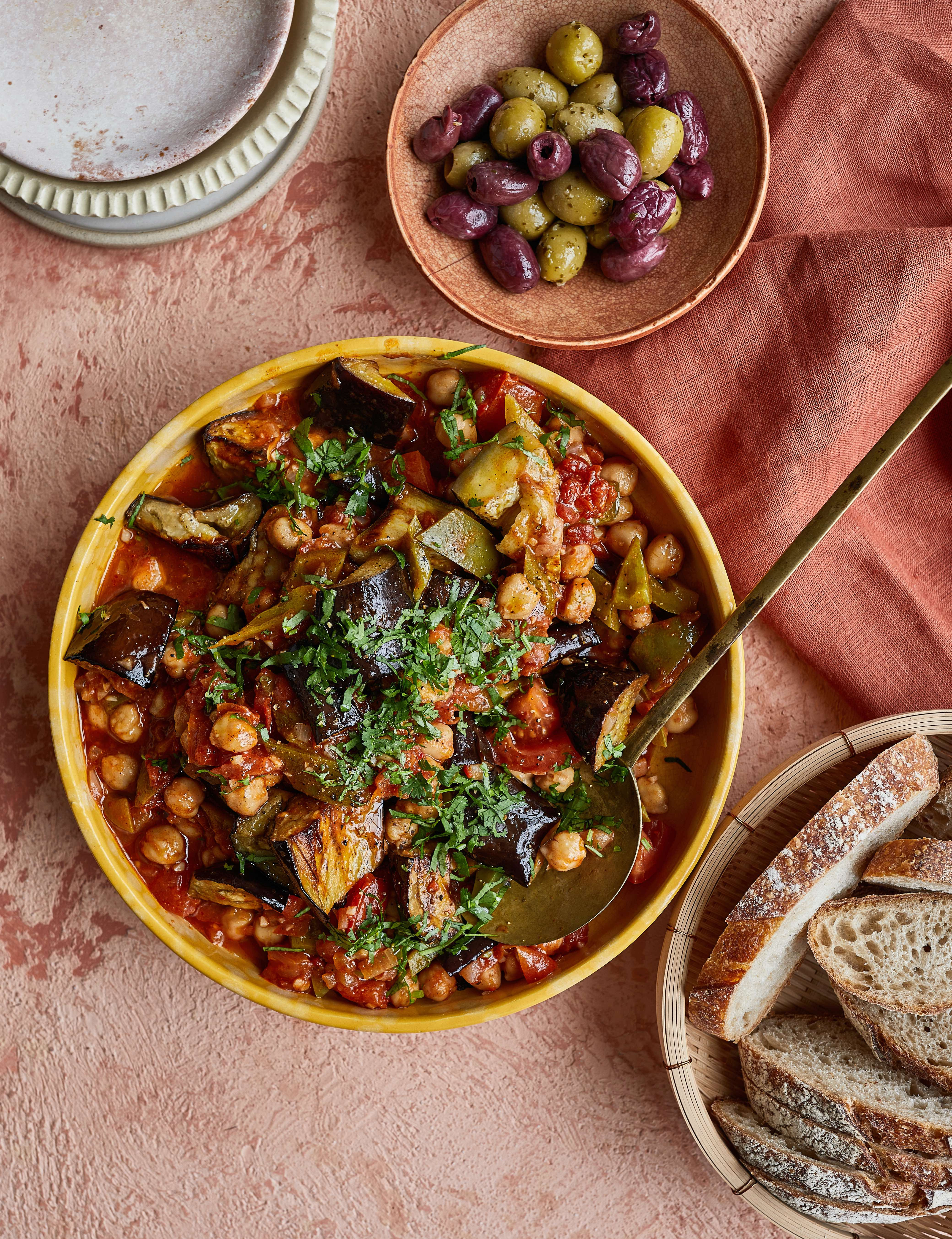 Yellow bowl with aubergine and chickpea stew, wooden bowl of mixed olives, bread basket, and empty plate on pink surface.
