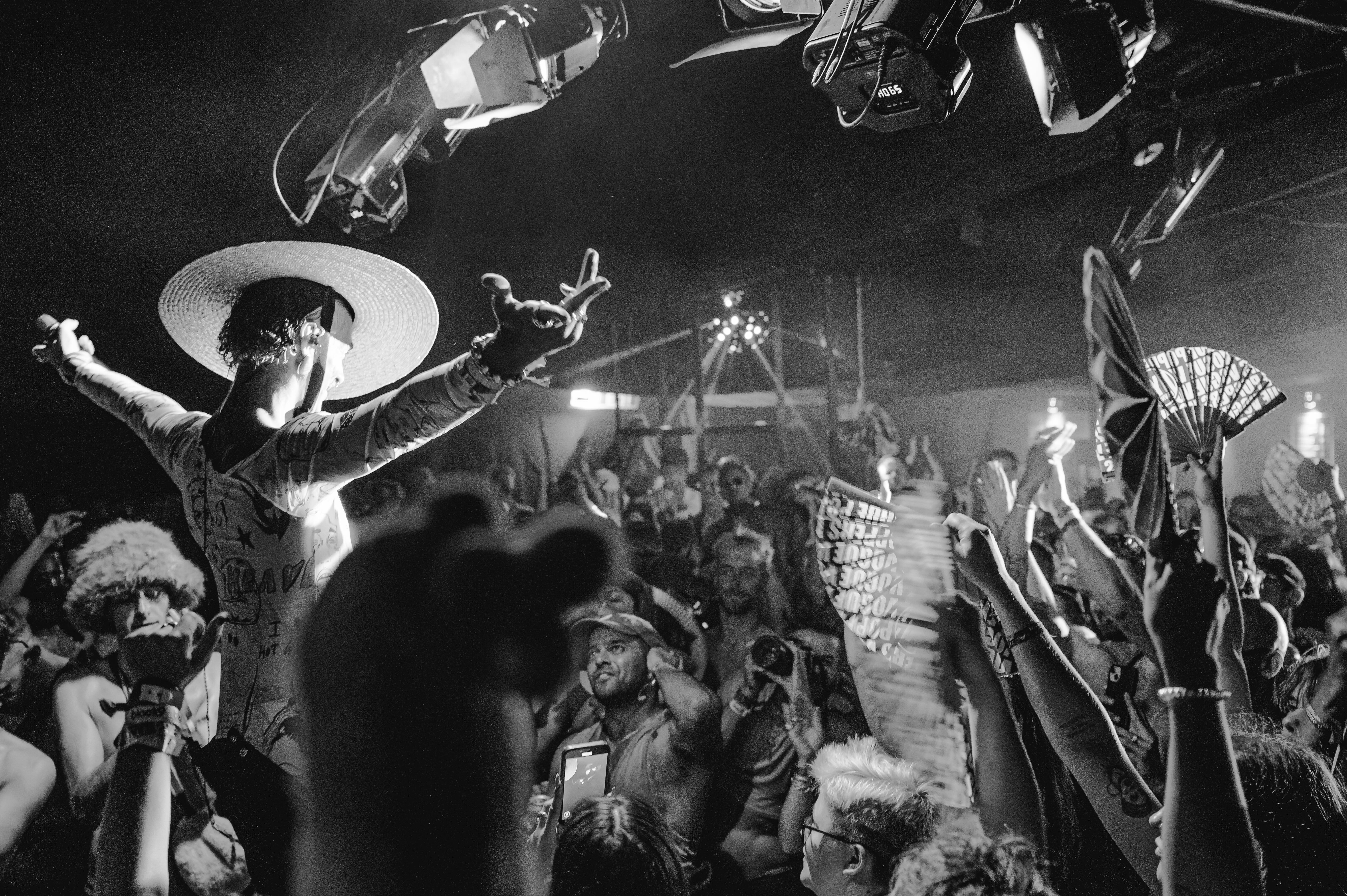 Black and white image of crowded indoor venue with performer in wide-brimmed hat on stage, audience below, dramatic lighting and shadows.