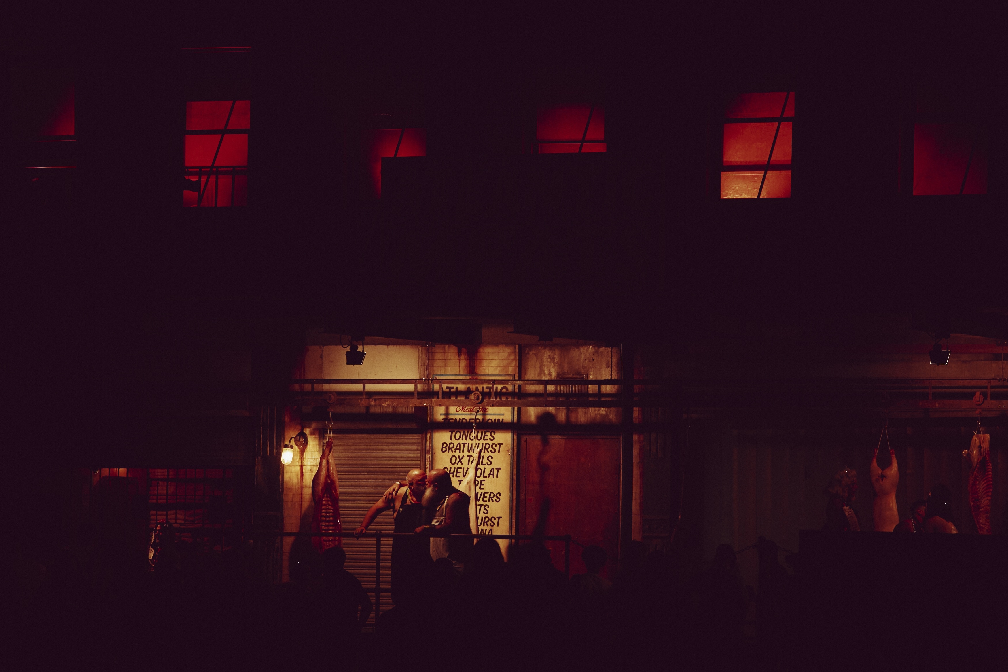Dark street scene with orange-lit shop front and glowing red windows in building above. People gathered outside illuminated storefront.