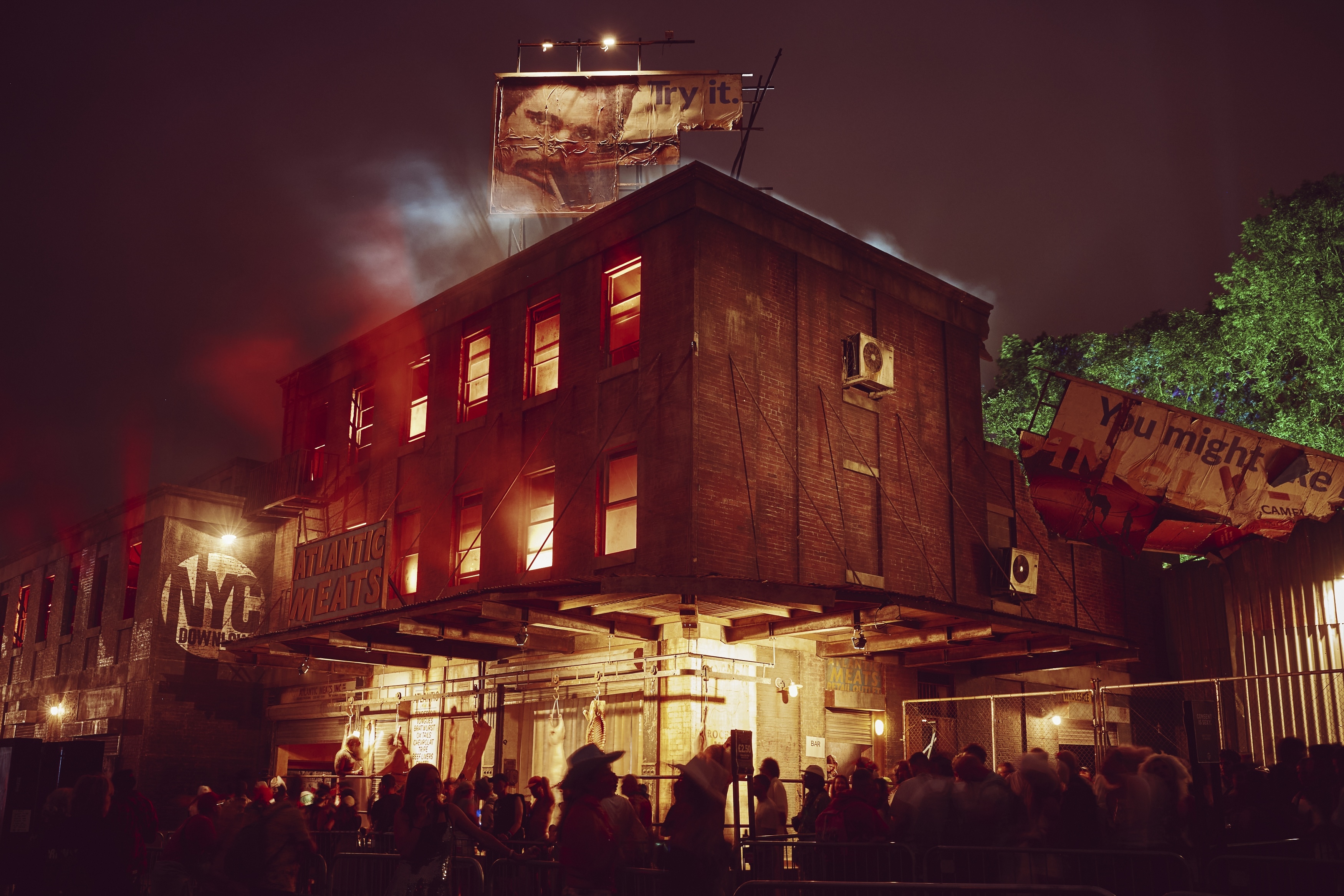 Industrial brick building at night with warm orange lighting from windows, crowd gathered below, dark purple sky, green foliage visible right side.