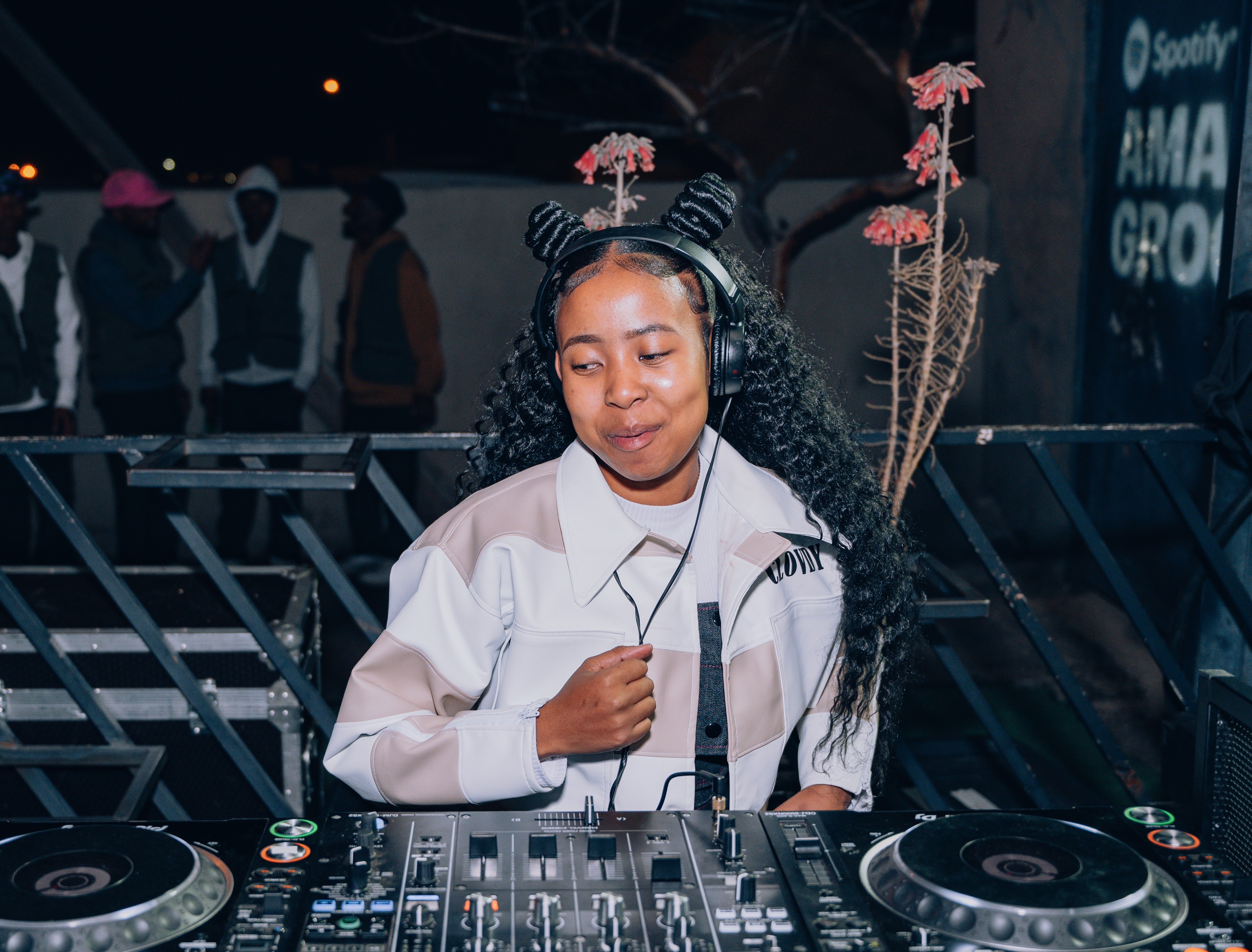 A young woman in a white shirt and headphones playing music on a DJ mixer at a night event.