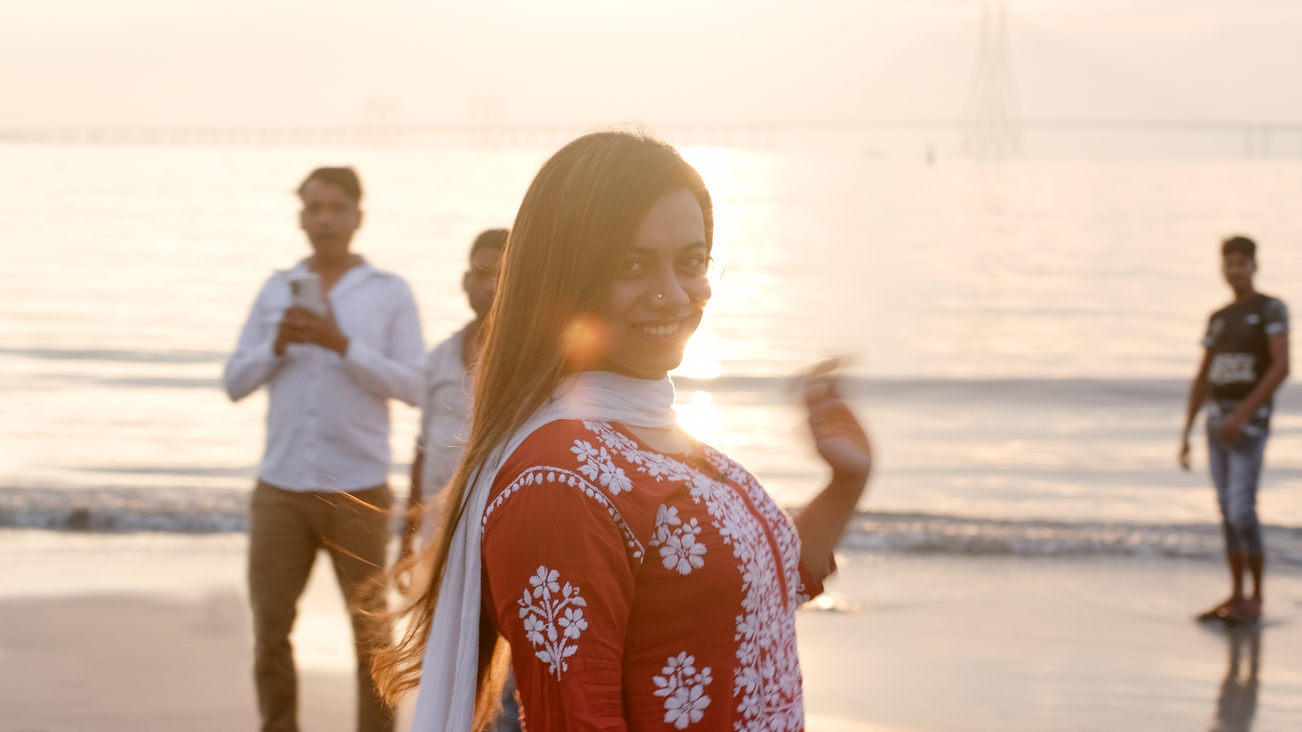 Woman in red patterned top smiling on beach at sunset, with three people in background near ocean waves.
