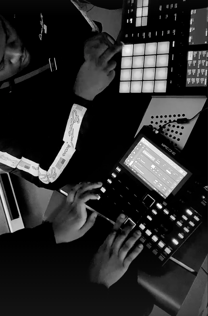 Person using electronic music equipment with grid controllers, laptop screen, and hands on control surfaces. Black and white image.